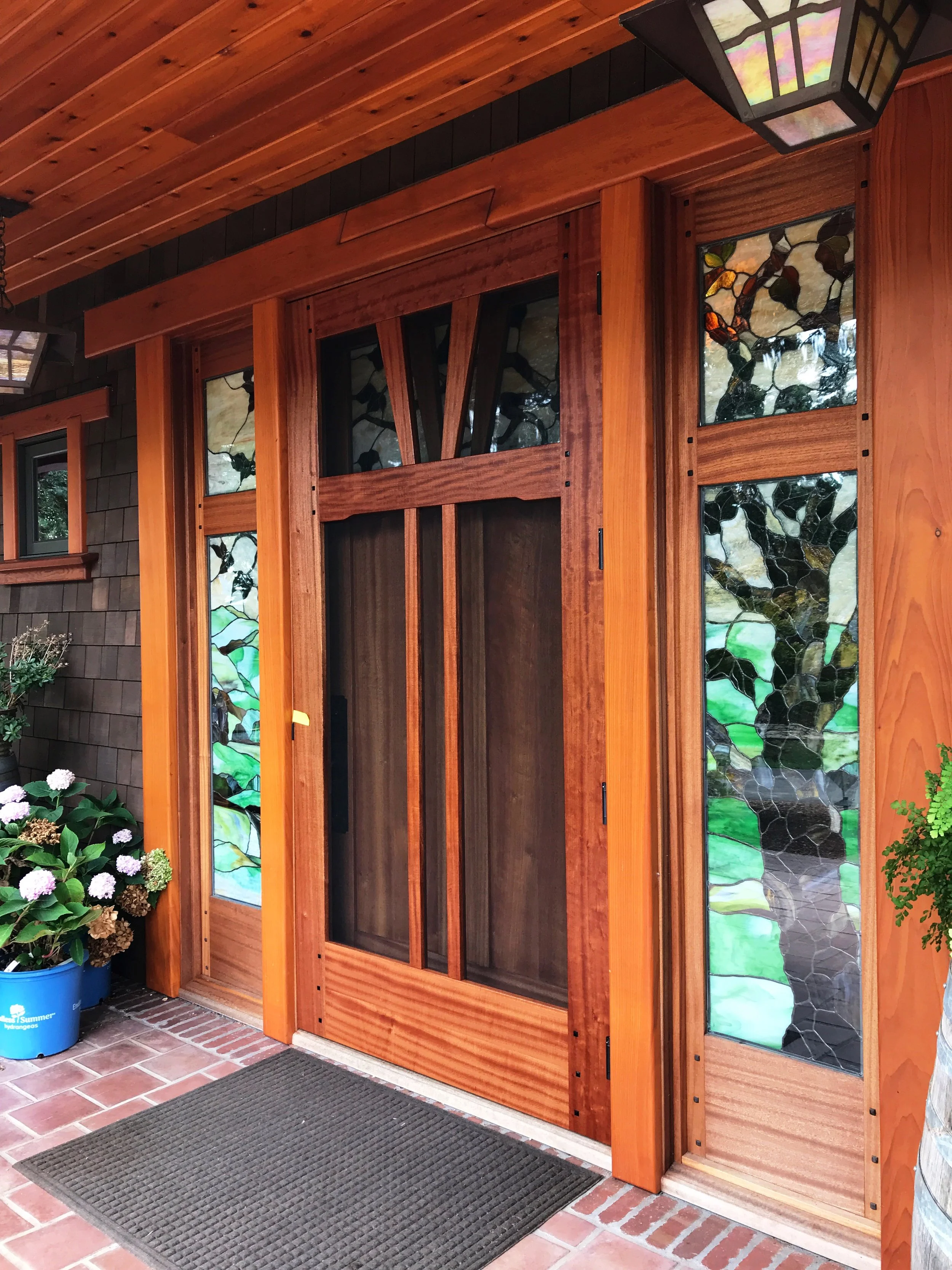 Ark Built Carpentry custom designed front door with wooden frame and stained glass side panels, black doormat on tile porch, potted hydrangea flowers on left.