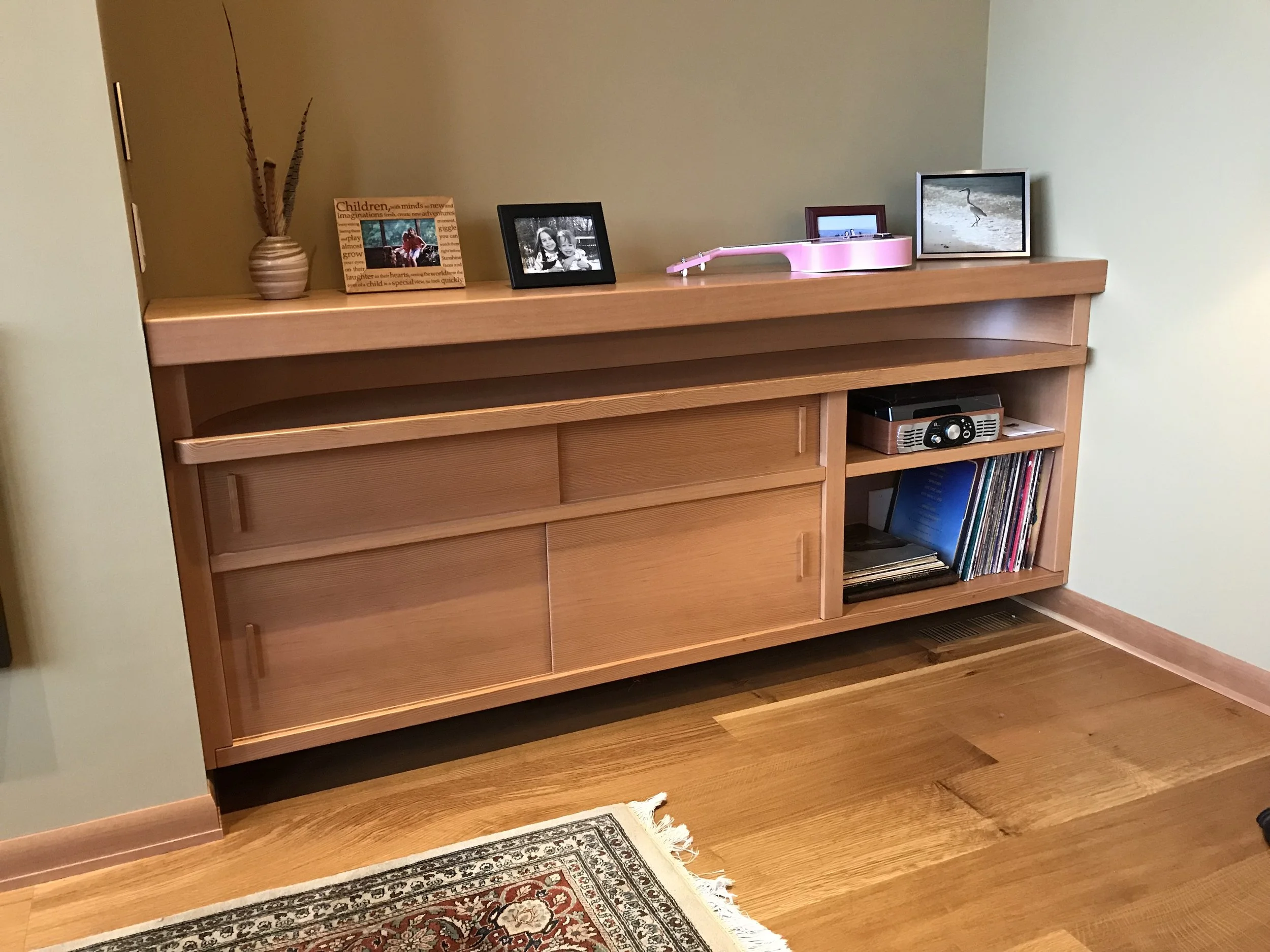 Ark Built Carpentry custom built wooden sideboard with framed photographs and a decorative vase with tall feathers on top, against a green wall, with a colorful rug on the hardwood floor in front.