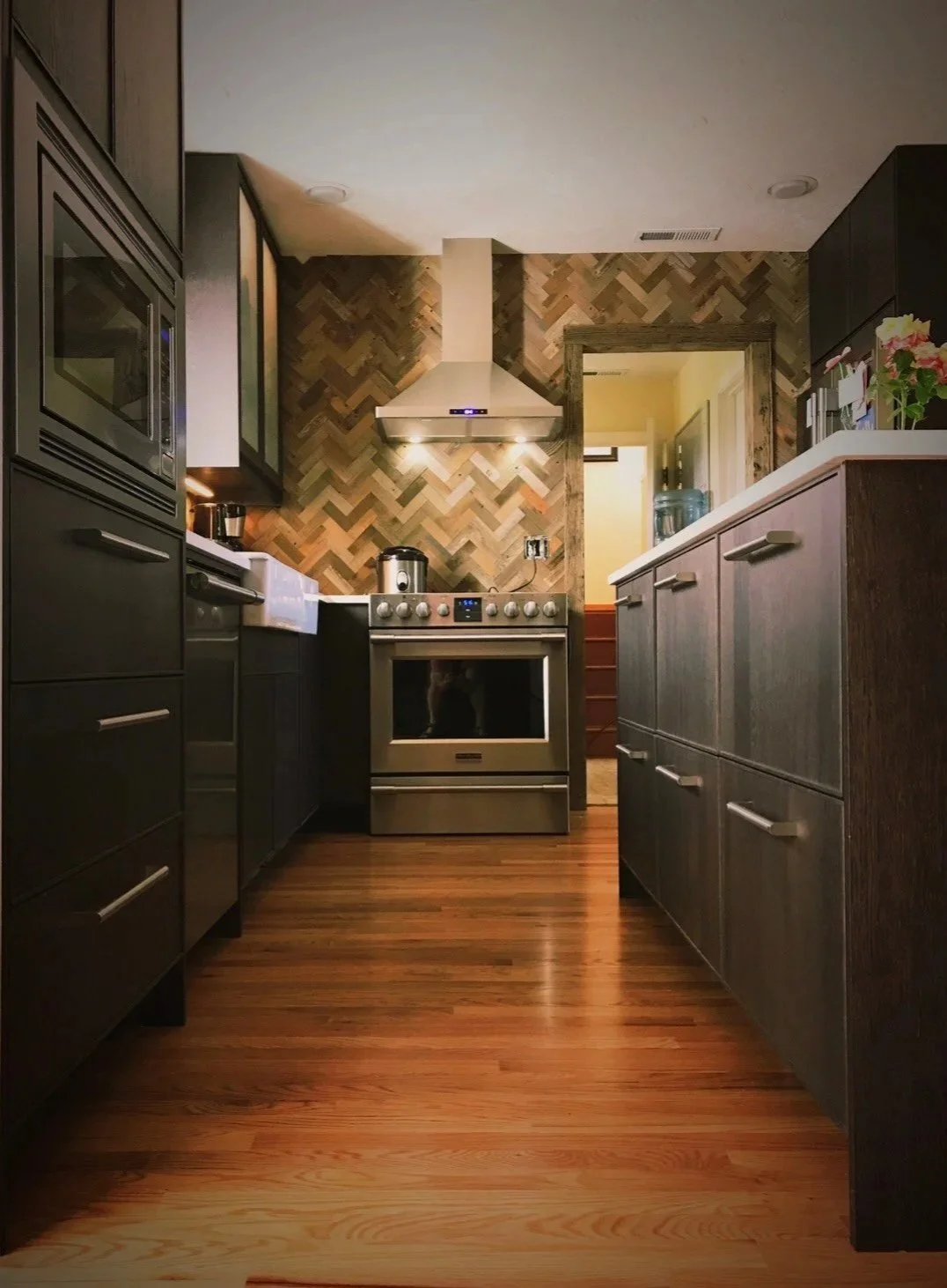 Ark Built Carpentry custom modern kitchen with dark cabinets, wood flooring, herringbone backsplash, stainless steel stove, range hood, and various appliances.
