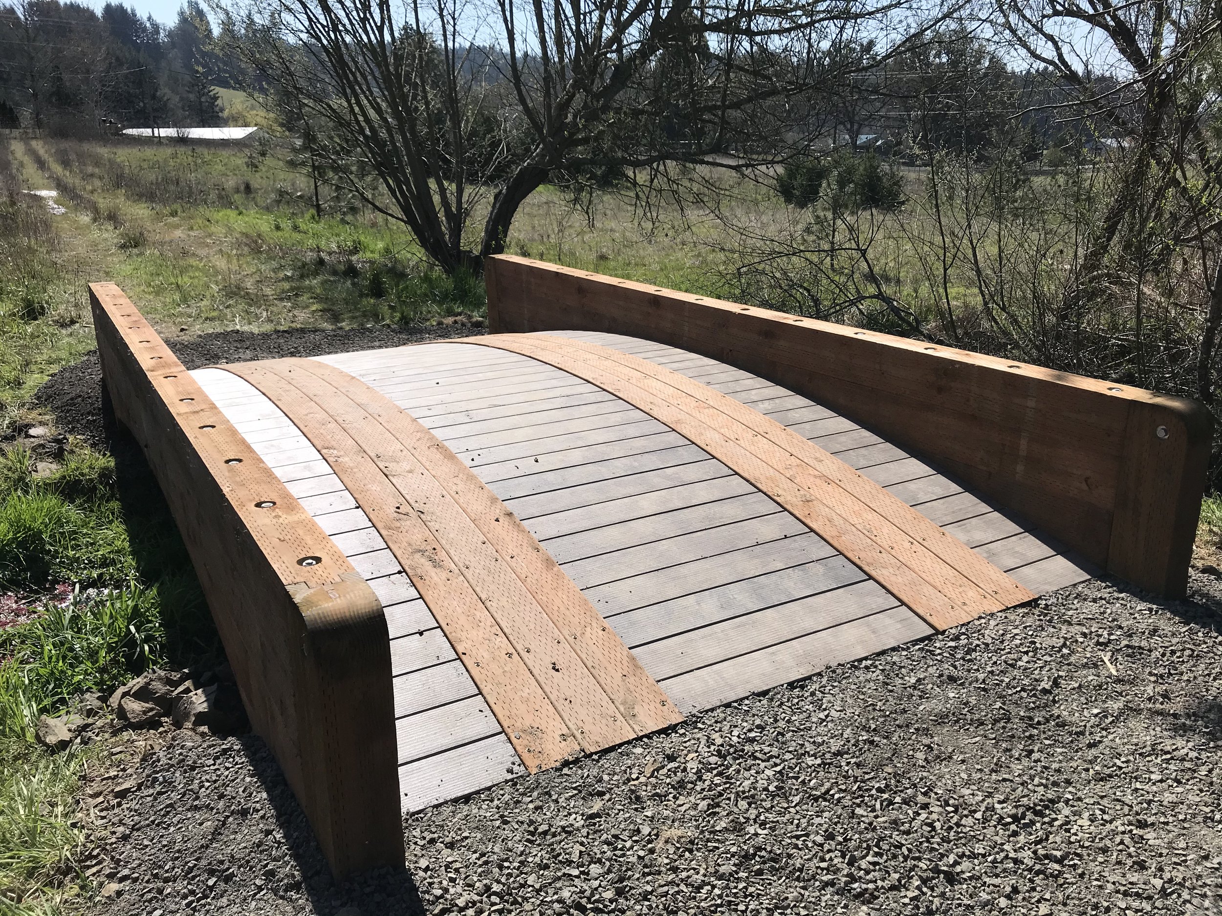 Ark Built Carpentry custom designed wooden bridge with a curved design is situated in a grassy outdoor area with trees and shrubs, under a clear sky.