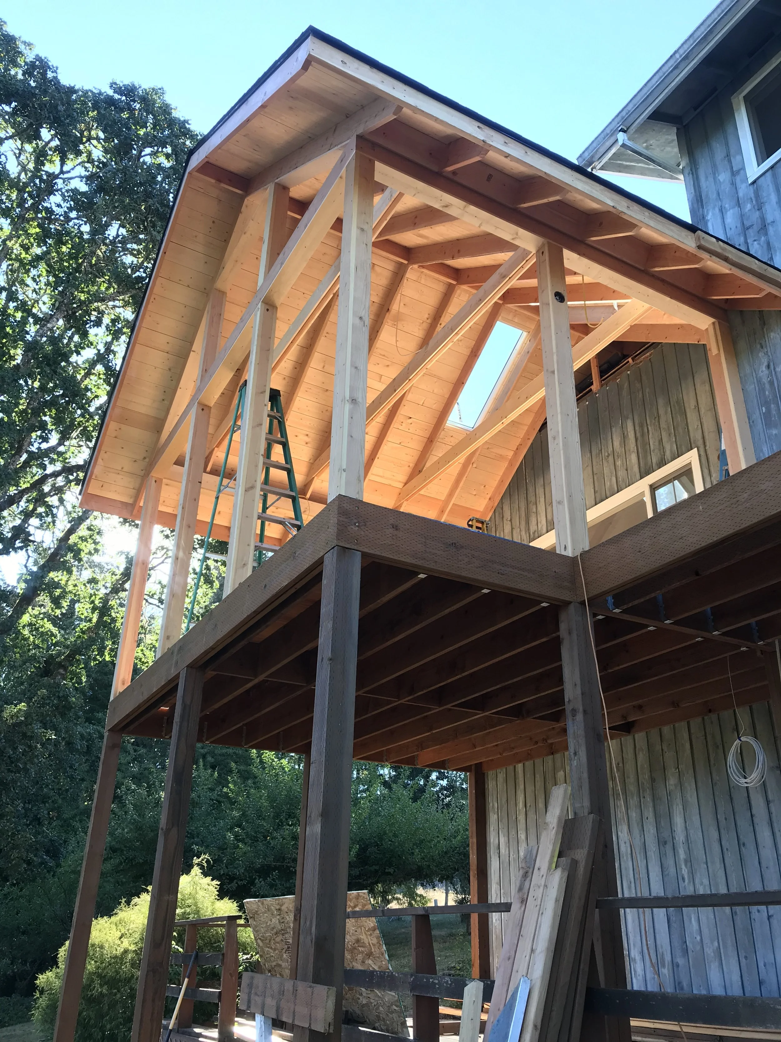 Ark Built Carpentry construction site of a house with framing for an exterior screened porch including vertical support posts, floor joists, and roof framing, with a ladder and building materials visible.