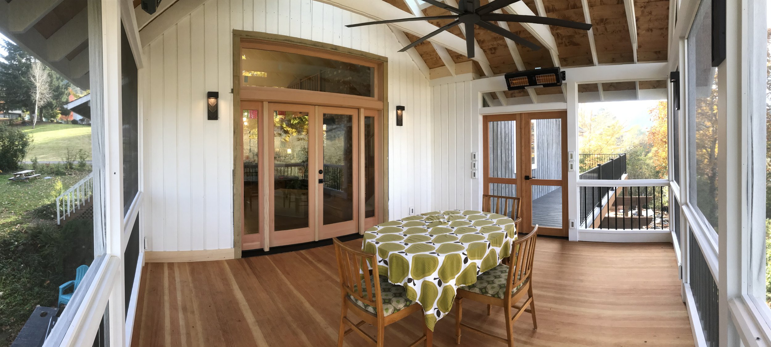 Ark Built Carpentry custom designed screened portch with white walls, large windows, and a wooden ceiling with a ceiling fan. There is a round table covered with a green and white patterned tablecloth, surrounded by four wooden chairs. 