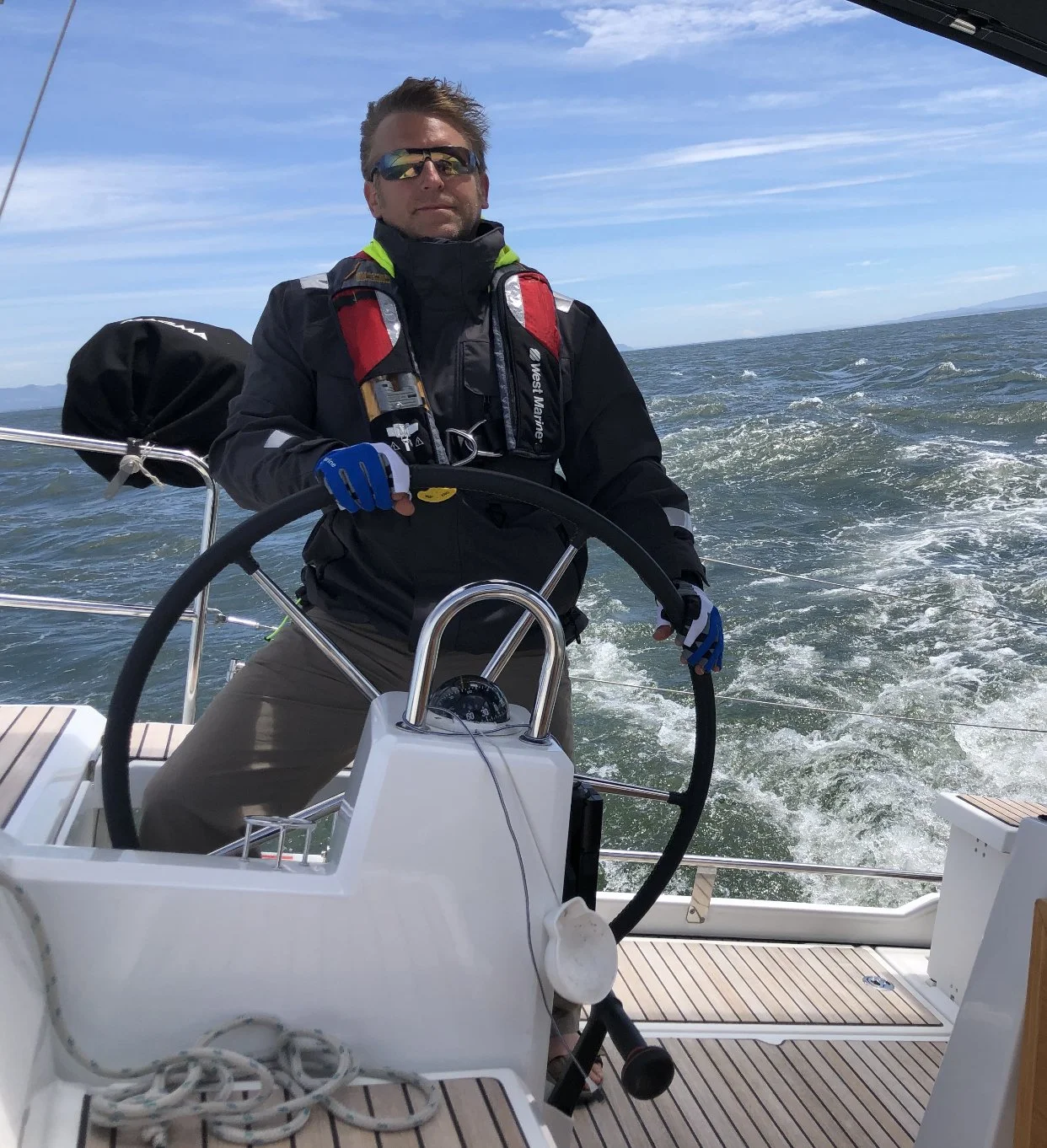 Ark Built Carpentry founder Matt Behnke steering a sailboat on the water, wearing sunglasses, a black jacket, and gloves, with open sea and blue sky in the background.