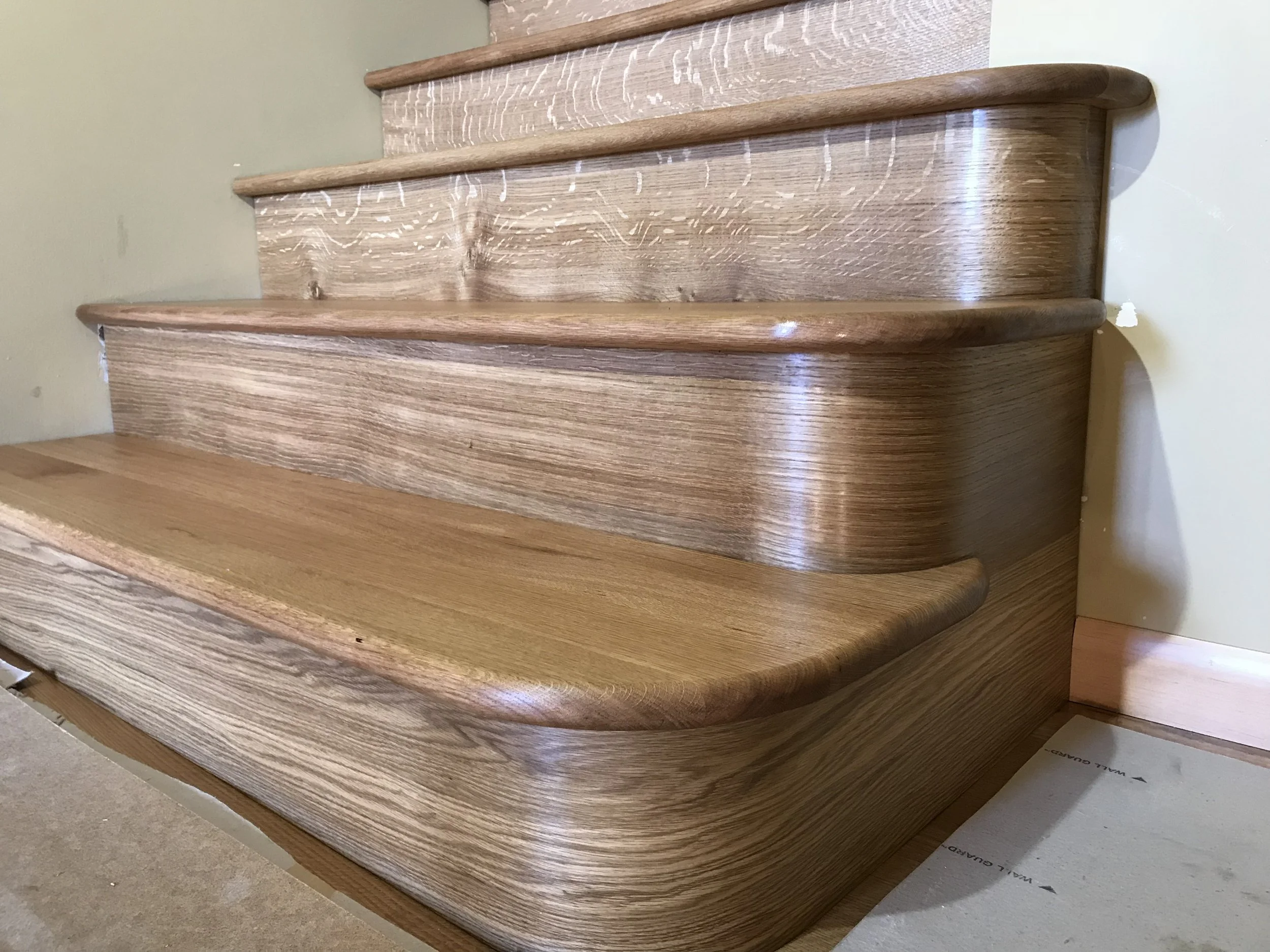 Ark Built Carpentry custom wooden staircase with rounded edges, with a beige wall behind. The staircase has three visible steps, each with a beautiful custom curved wood and smooth, polished finish.