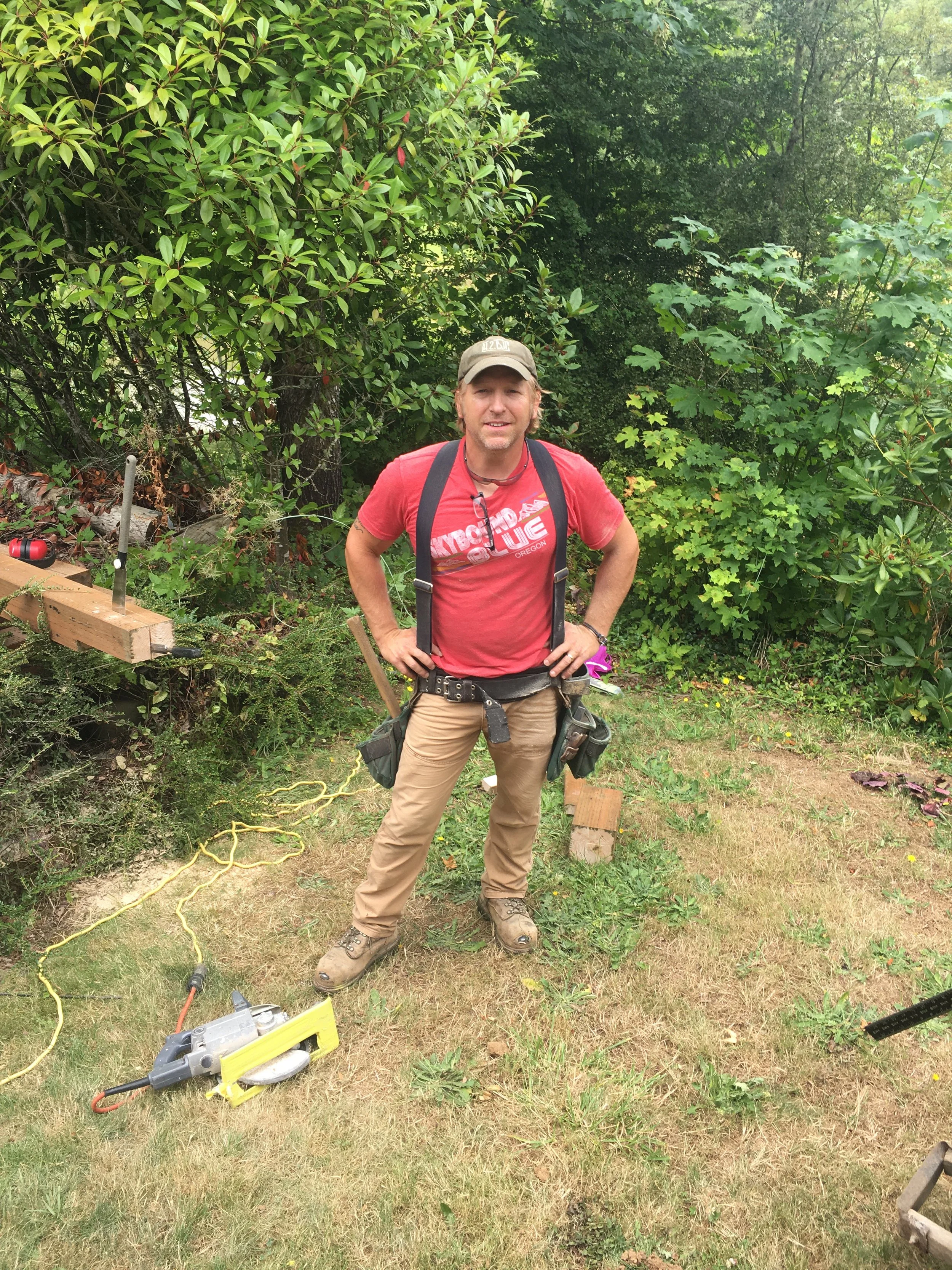 Ark Built Carpentry founder Matt Behnke stands outdoors on grass, surrounded by bushes and trees, wearing a red Skybound Blue t-shirt, tan pants, work boots, a cap, and tools attached to his belt, with woodworking tools nearby.
