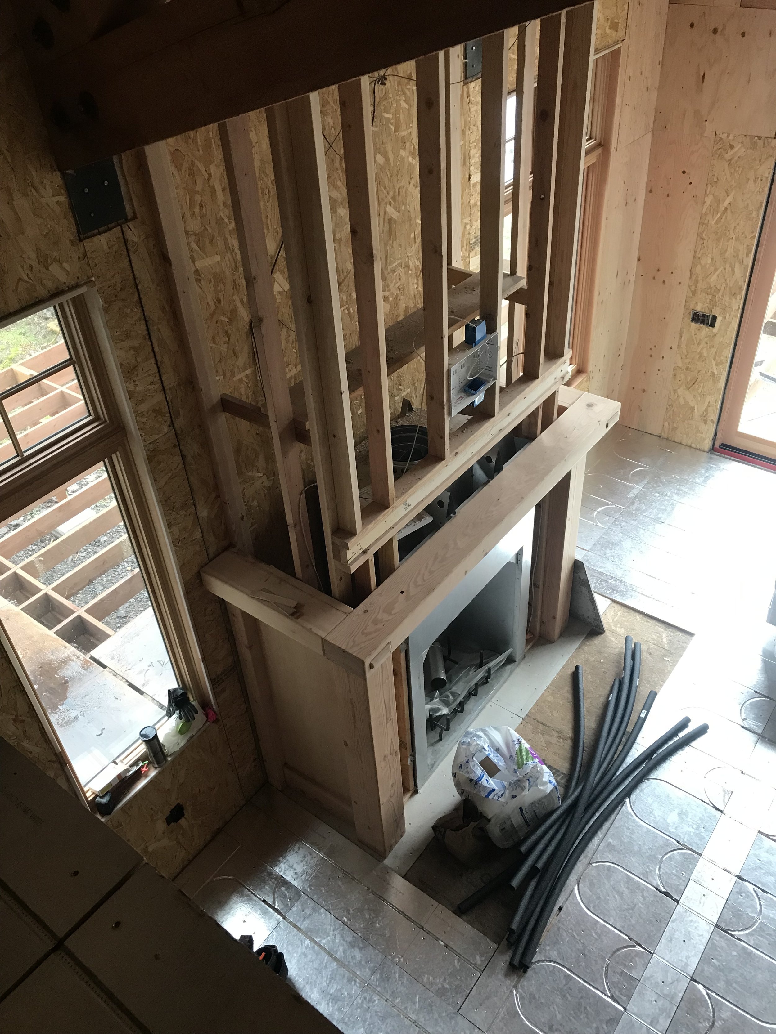 Ark Built Carpentry custom designed fireplace mantel Interior view of a house under construction with wooden framing, a window, and construction tools and materials on the floor.