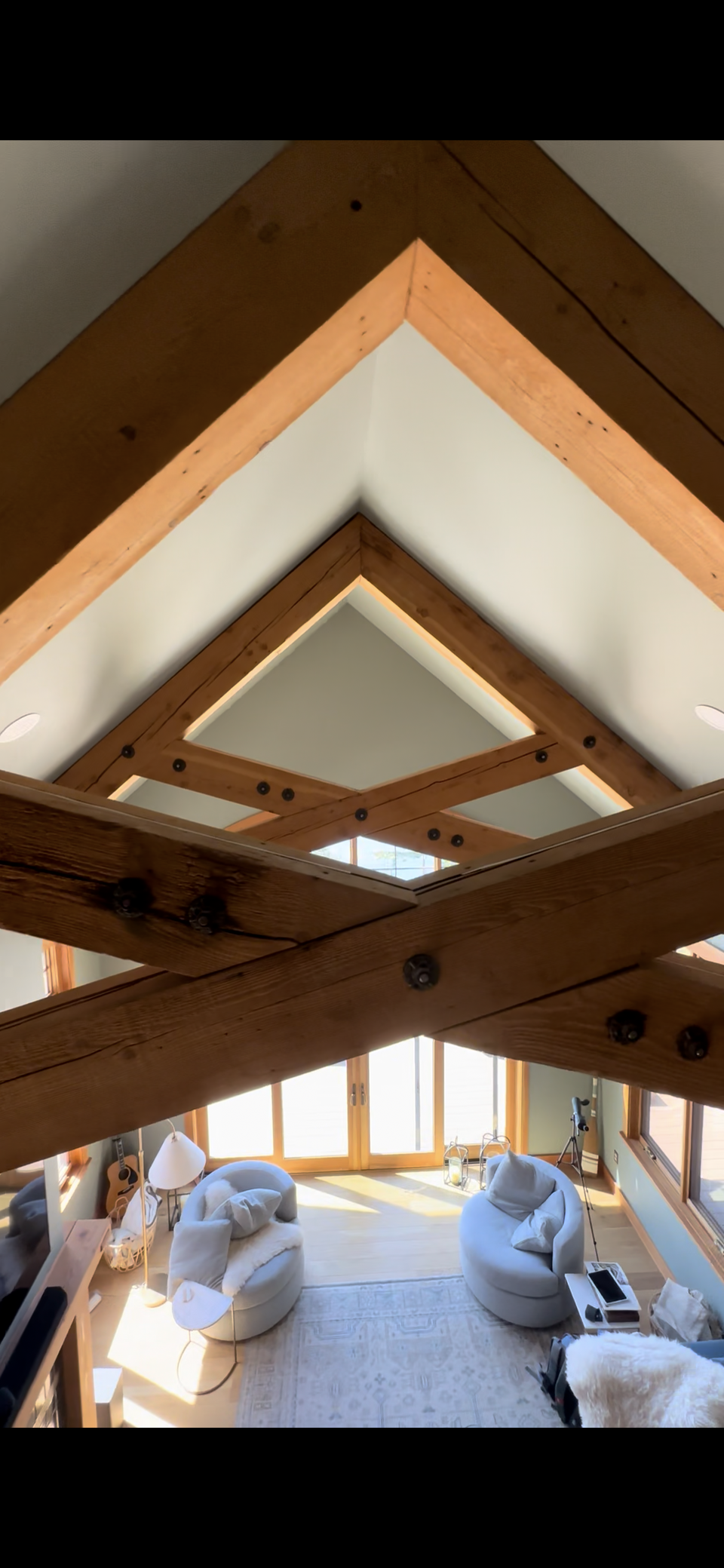 Ark Built Carpentry custom beams Living room with large windows, two round white chairs, a white sofa with a blanket, an acoustic guitar, a floor lamp, and a telescope, viewed from a loft with exposed wooden beams and ceiling.