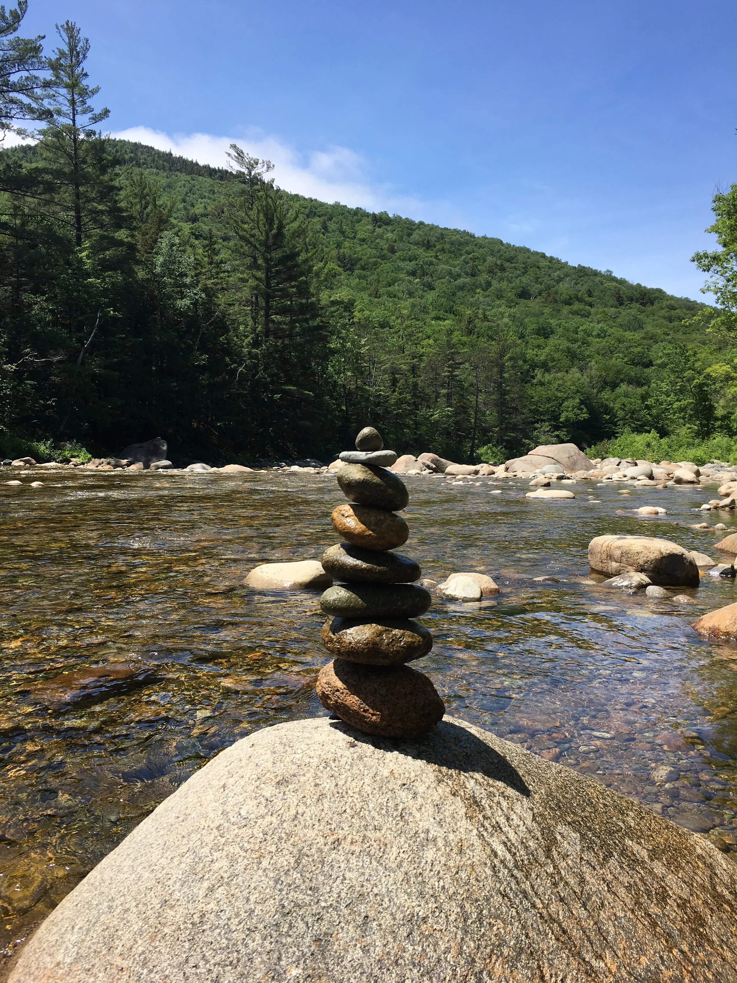 Piles of stacked rocks in a shallow river with a forested mountain and blue sky in the background.