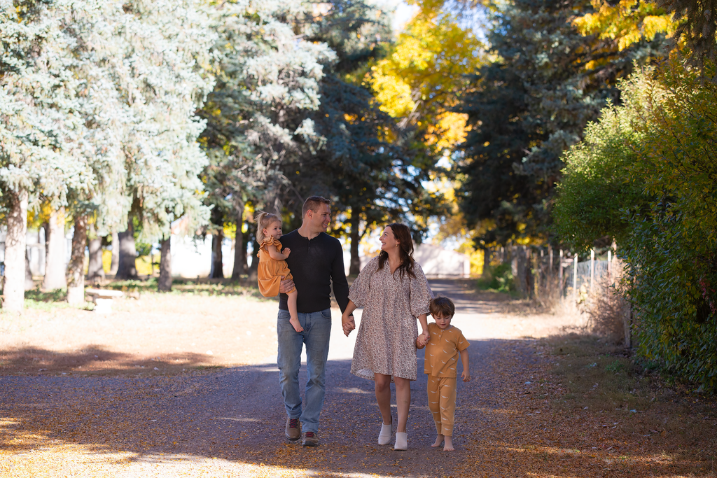 A family of four walking on a tree-lined dirt path during autumn, holding hands and enjoying each other's company.