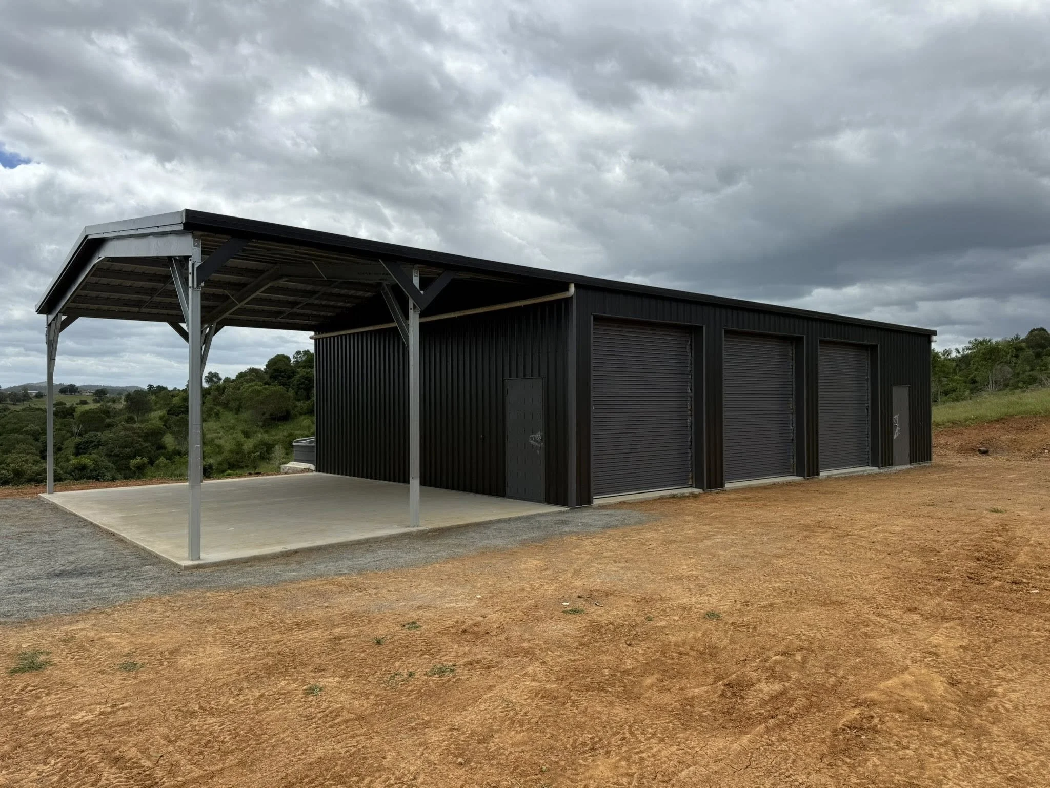 A metal storage shed with three roll-up doors and a side door, situated on a concrete pad with a covered area supported by metal posts, located in a rural area with dirt ground and green landscape in the background under gray cloudy sky.