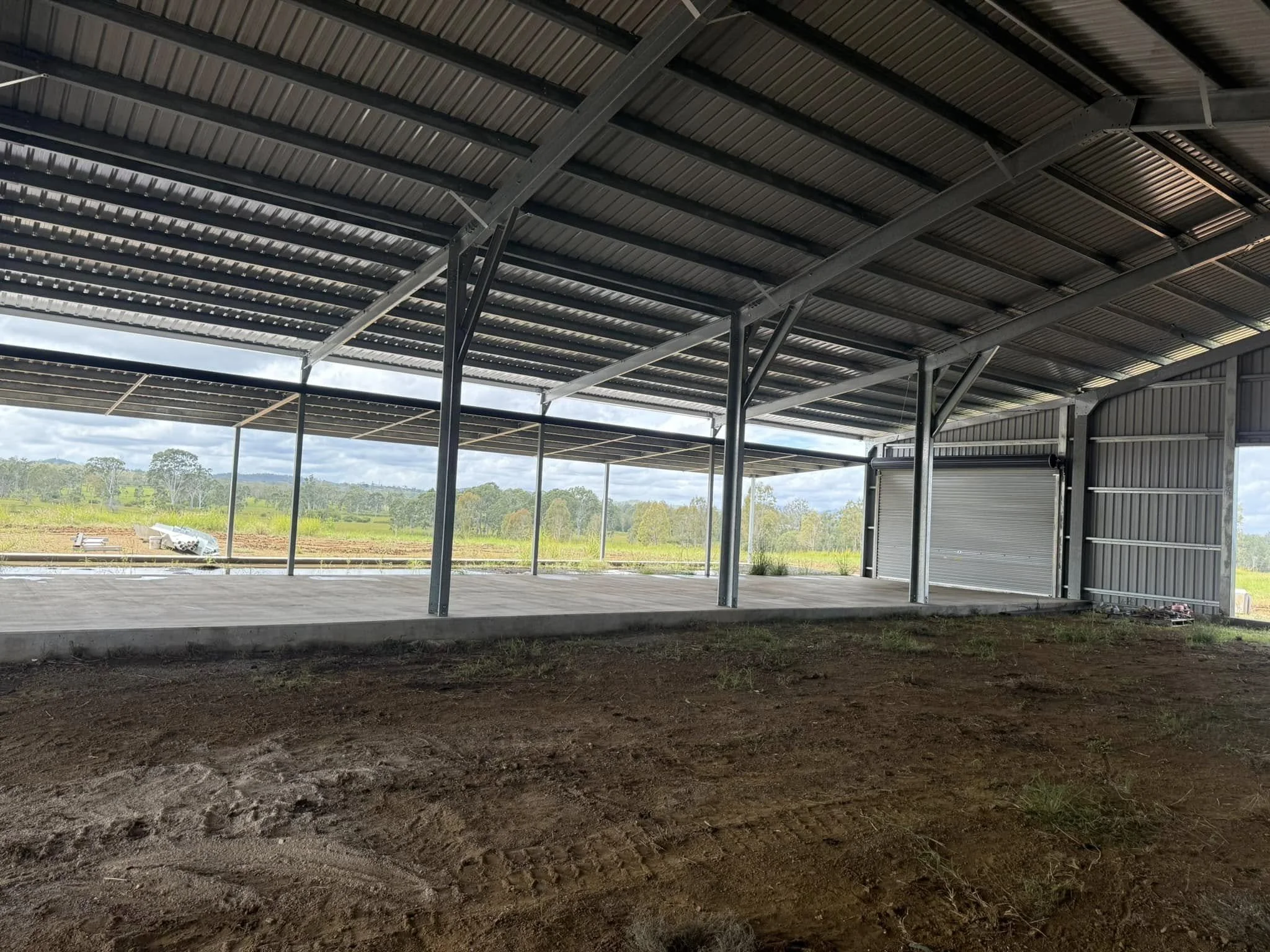 Arena style metal building with a concrete floor, open sides, and a rolled-up garage door, overlooking a rural landscape with fields and trees.