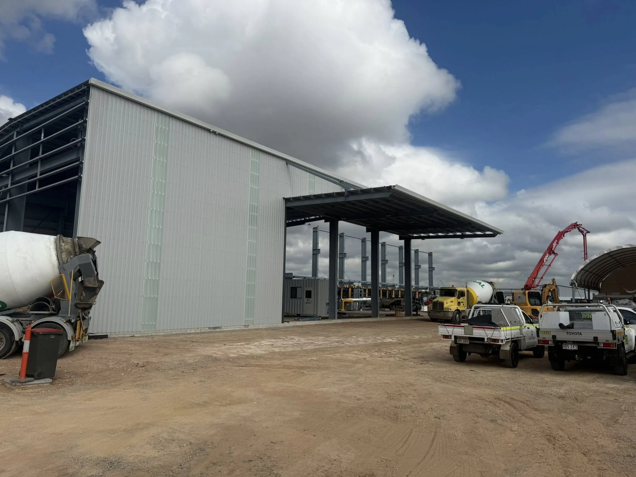 Construction site with a large metal building, concrete mixer truck, construction vehicles, and a crane under a partly cloudy sky.