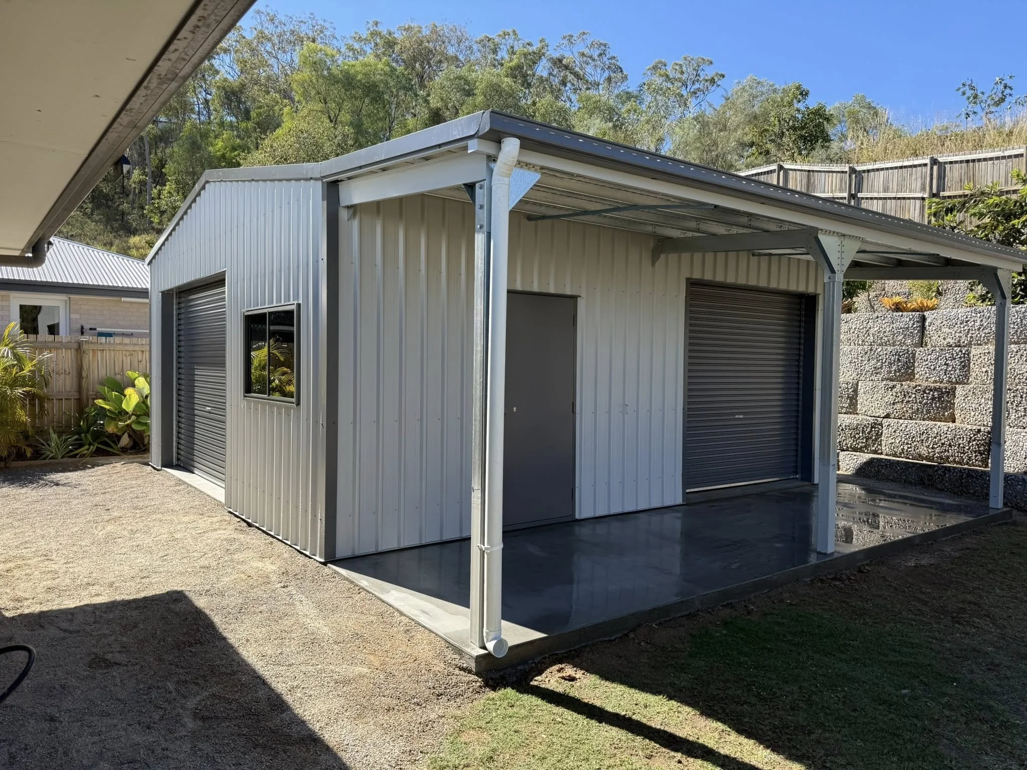 A metal garage with two roller doors, a window, and a door under a covered extension, set in a yard with a wooden fence and trees in the background.