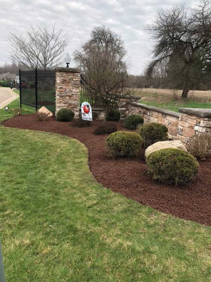 Landscape view of a landscaped yard with a stone wall, black metal gate, and mulched flower beds with shrubs and rocks, under an overcast sky.