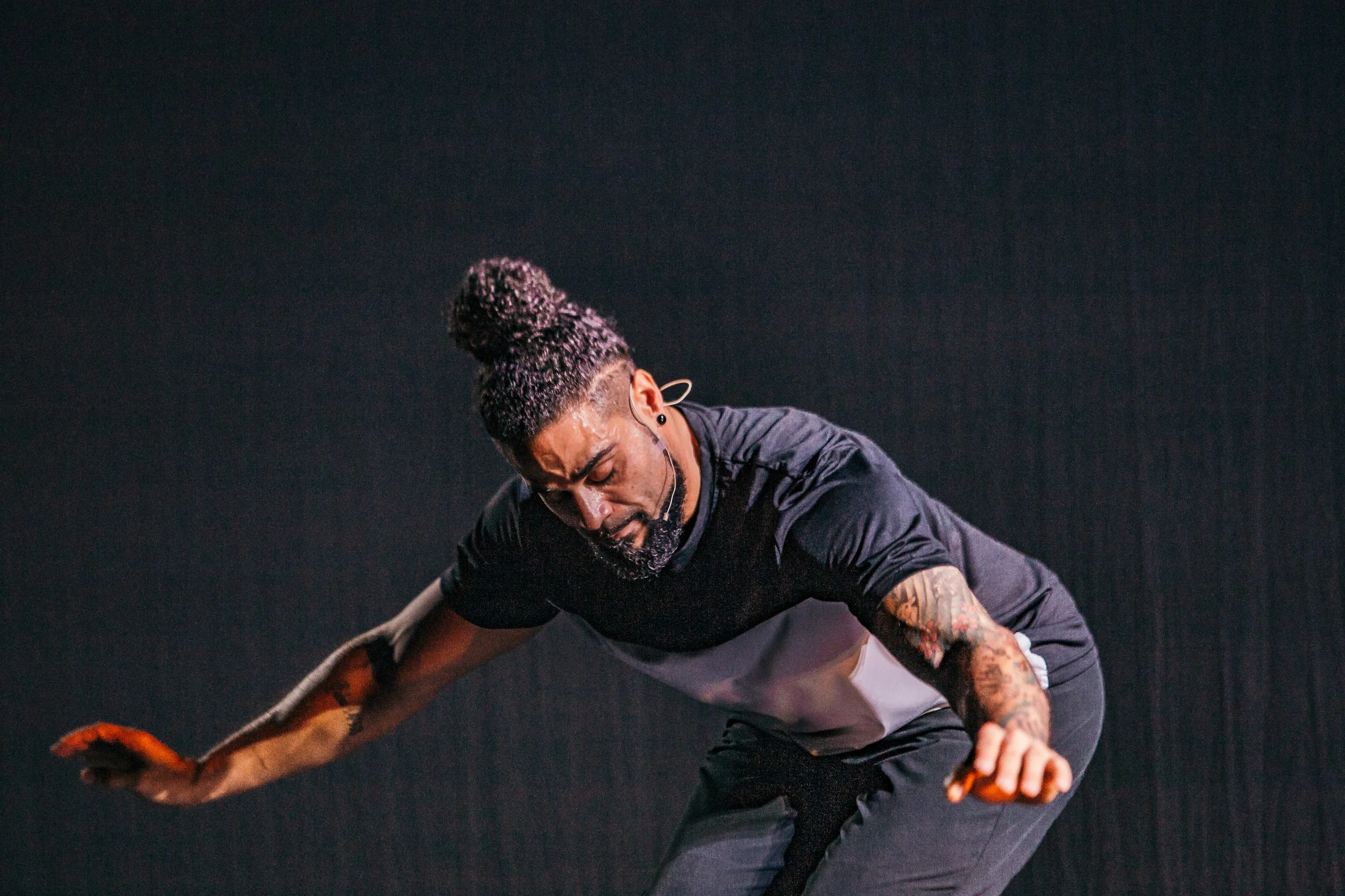 L.D., with tattoos on his arms, a man bun hairstyle, and a beard, wears a black and white athletic shirt and pants, leaning forward with his eyes closed and a strained expression, against a plain black background.