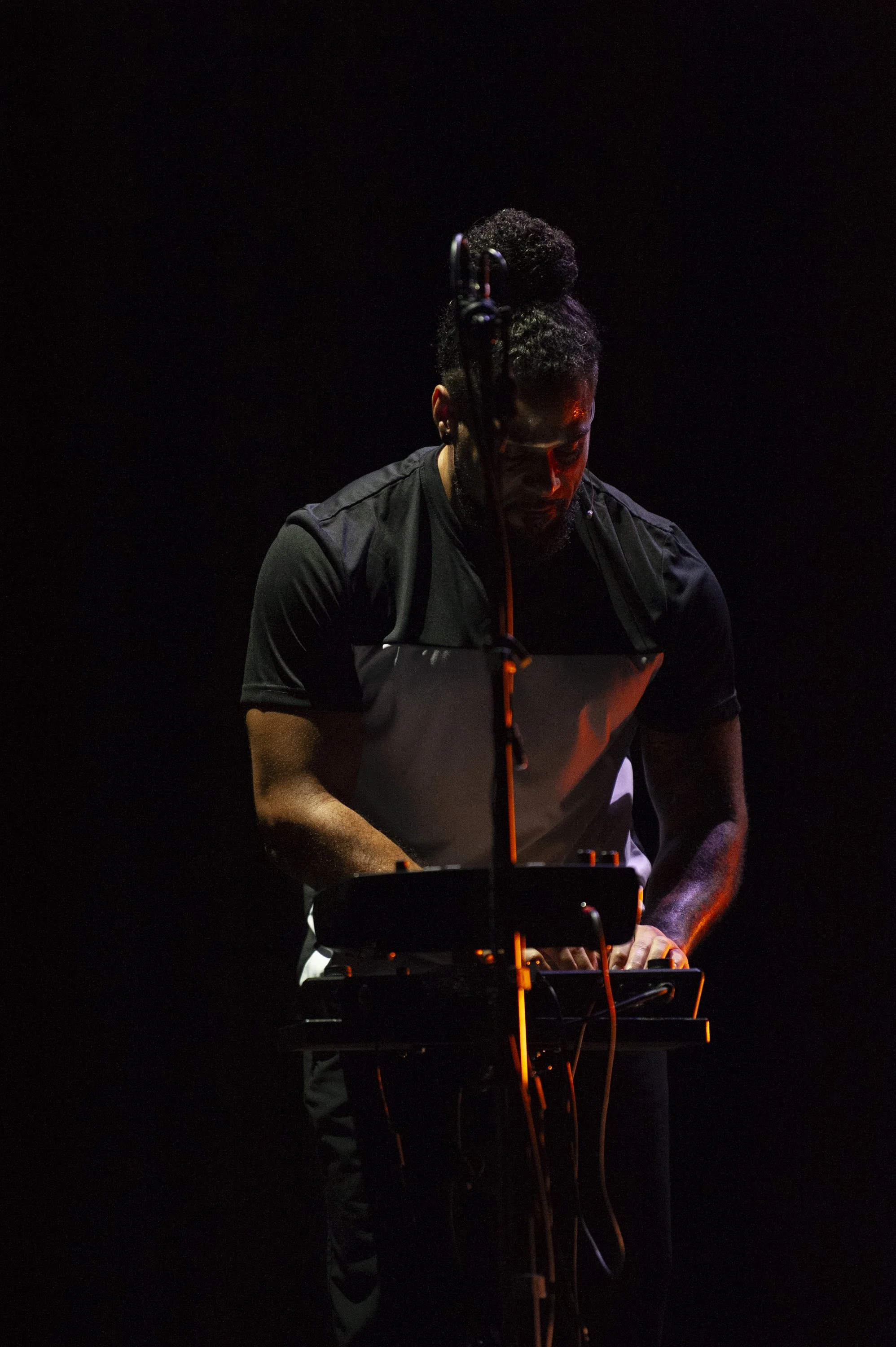 A man performing on stage with a keyboard, with a dark background, wearing a black shirt, illuminated by stage lights.