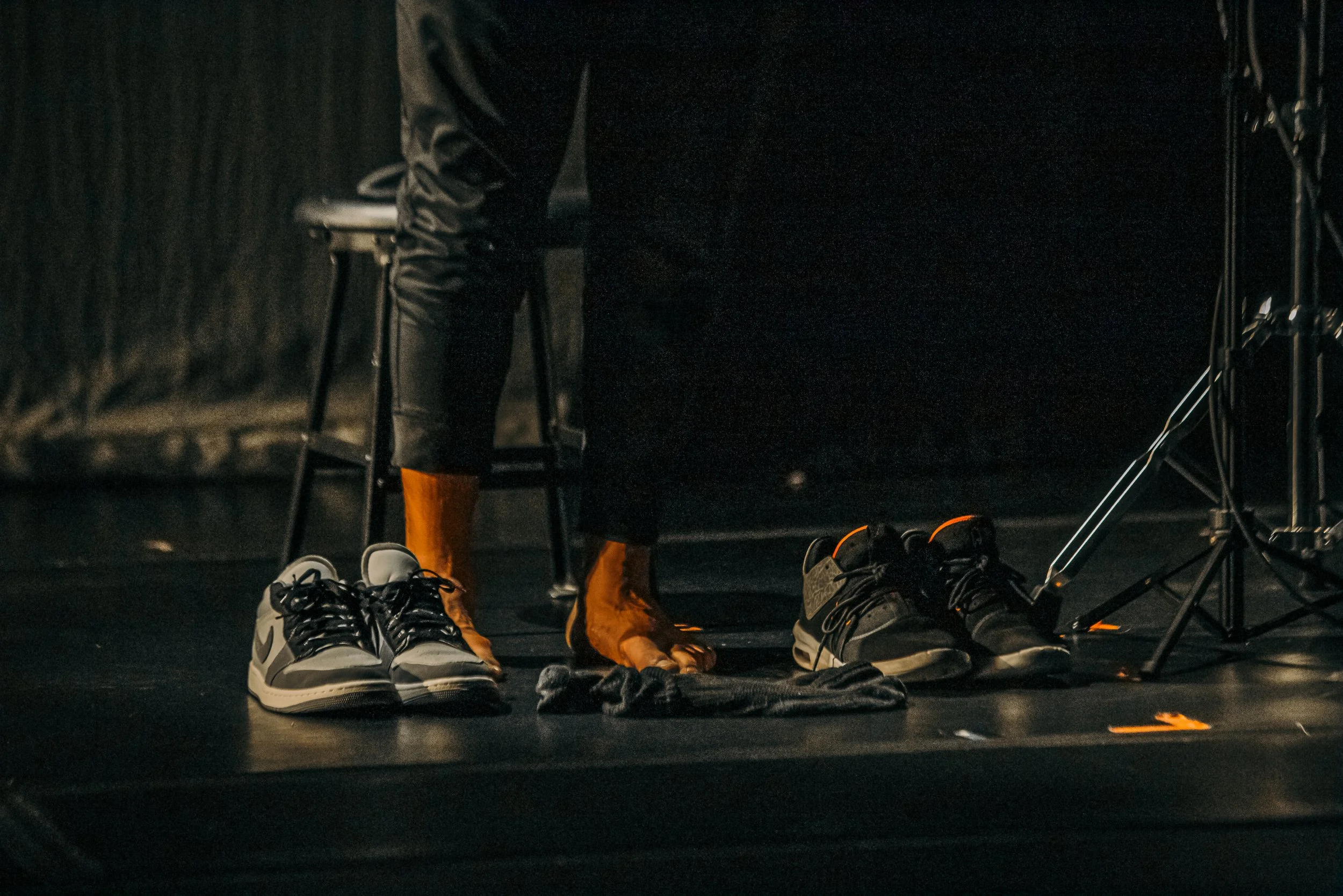 Footwear including sneakers, orange rain boots, and a pair of black shoes on a dark stage with a person partially visible wearing black pants and a black jacket.