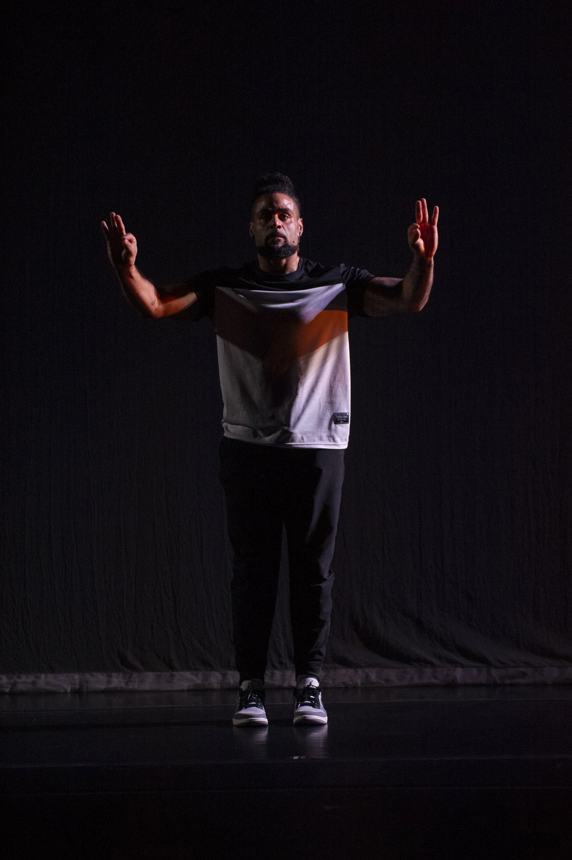 A man standing on stage with both arms raised, wearing a black and white t-shirt, black pants, and sneakers, against a dark background.