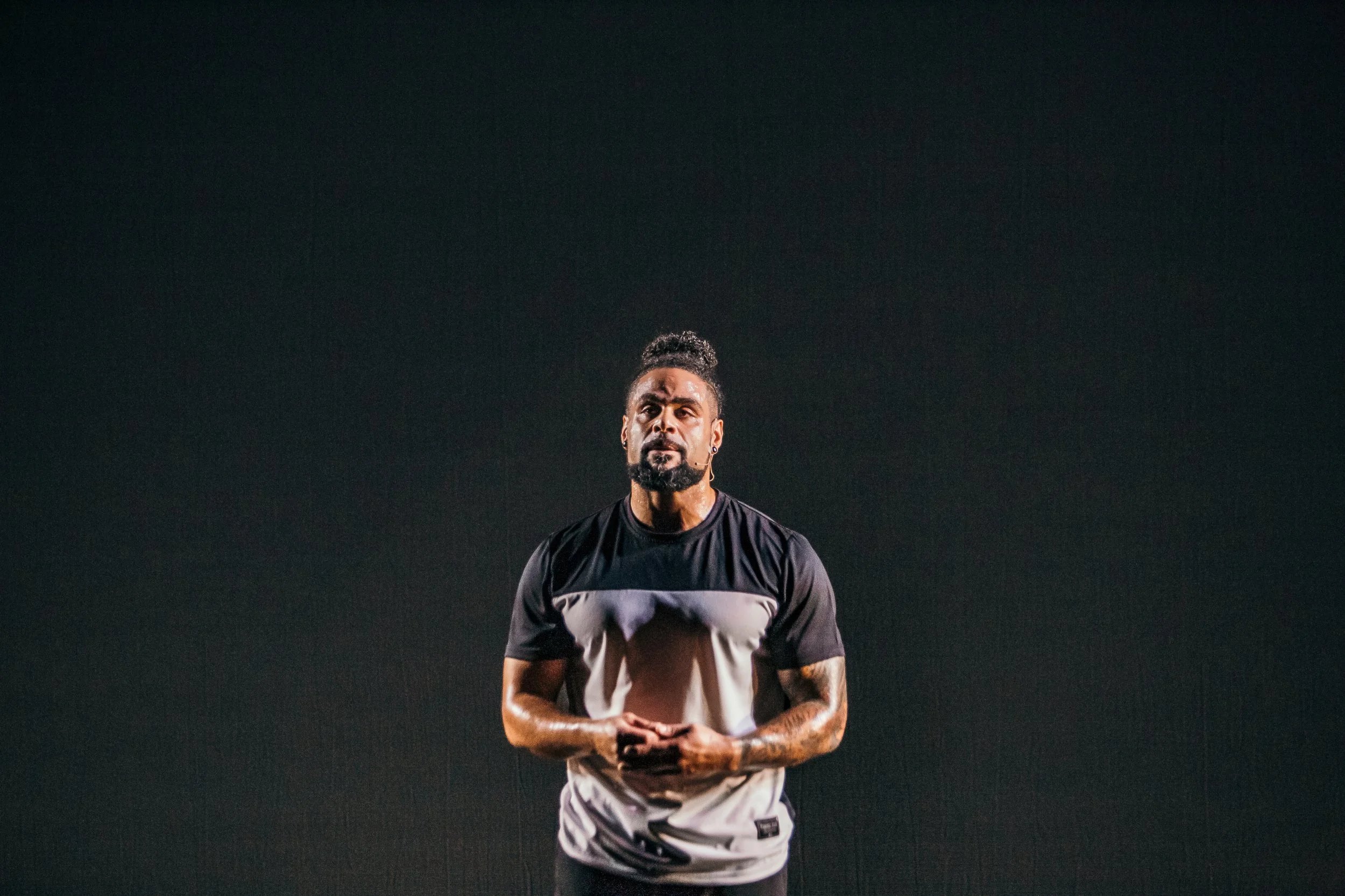 A man stands in front of a dark, empty background, wearing a black and white t-shirt with tattoos on his arms, and has a beard and styled hair.