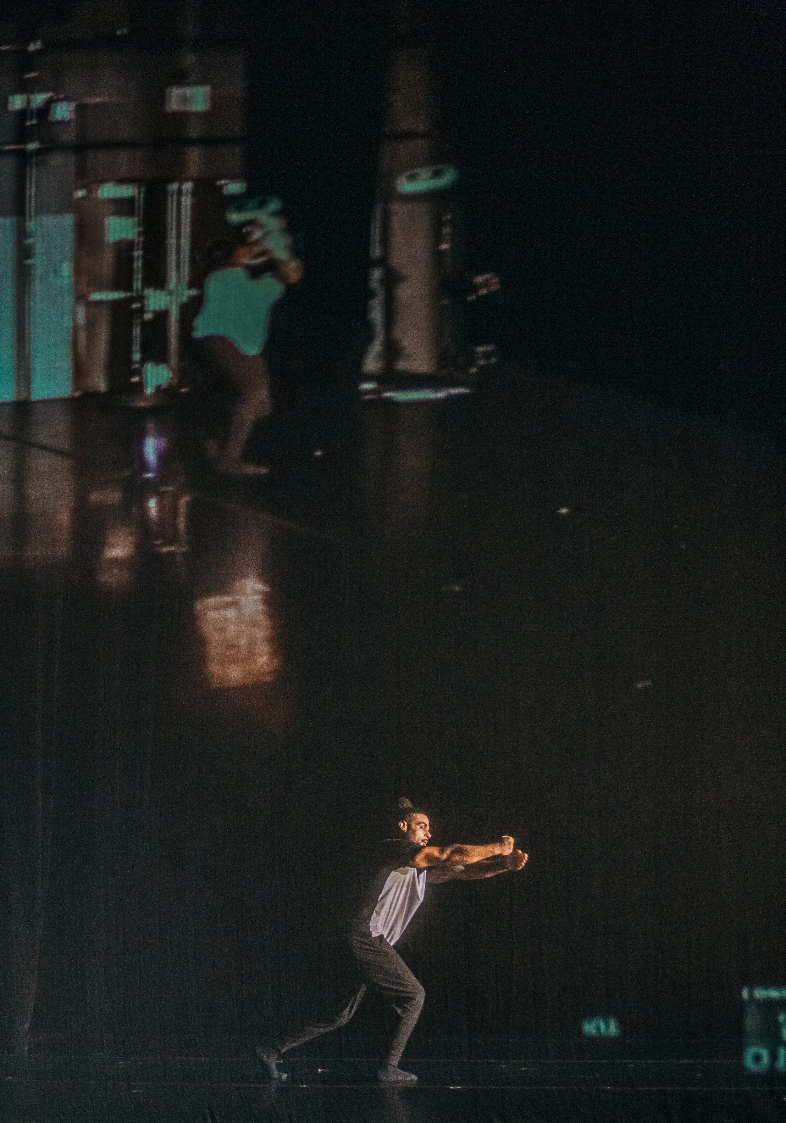 A man in a black shirt and pants is in a dance studio, performing a squat with arms extended forward.
