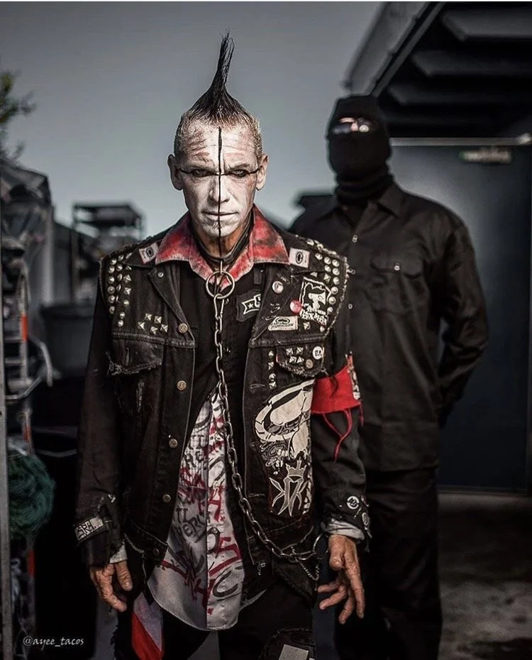 A man with a mohawk hairstyle, white black paint,  on his face and  dressed in a black studded denim jacket with patches and graffiti,  with a chain hanging from his neck, standing in front of a man in dark clothing with his face covered.