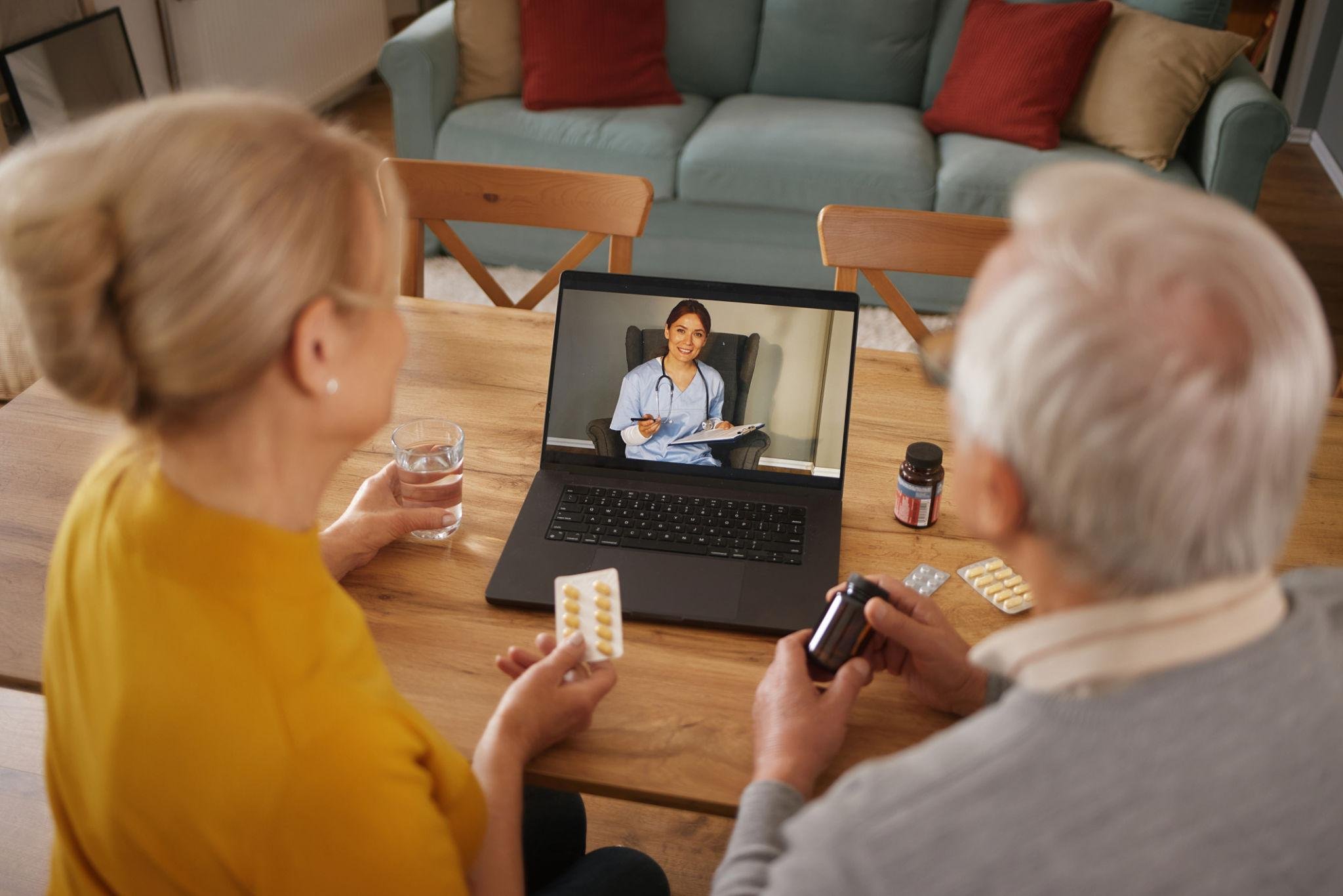 An elderly couple sitting at a dining table, having a telehealth consultation with a health provider on a laptop; the woman holds yellow pills in a blister pack, and the man holds a pill bottle and a glass of water.