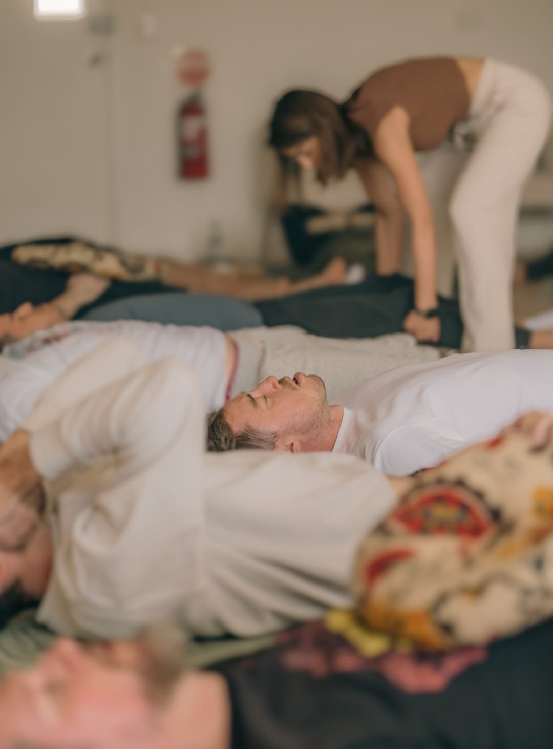 People lying down on their backs with eyes closed, while a woman in brown top and white pants leans over one person, in what appears to be a group meditation or relaxation session.