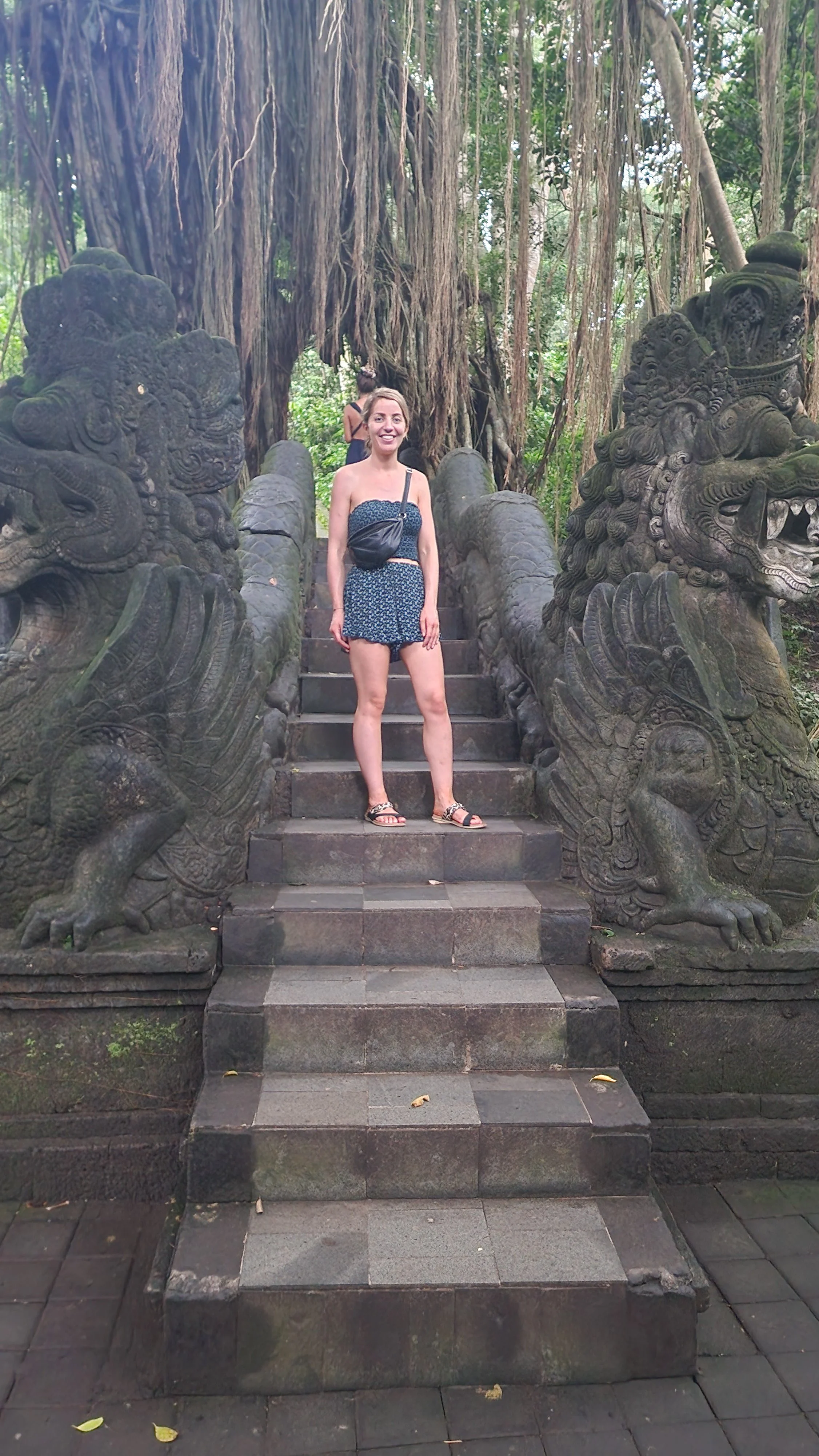 A woman standing on stone steps flanked by large carved stone sculptures of mythical creatures, with a large tree and greenery in the background.