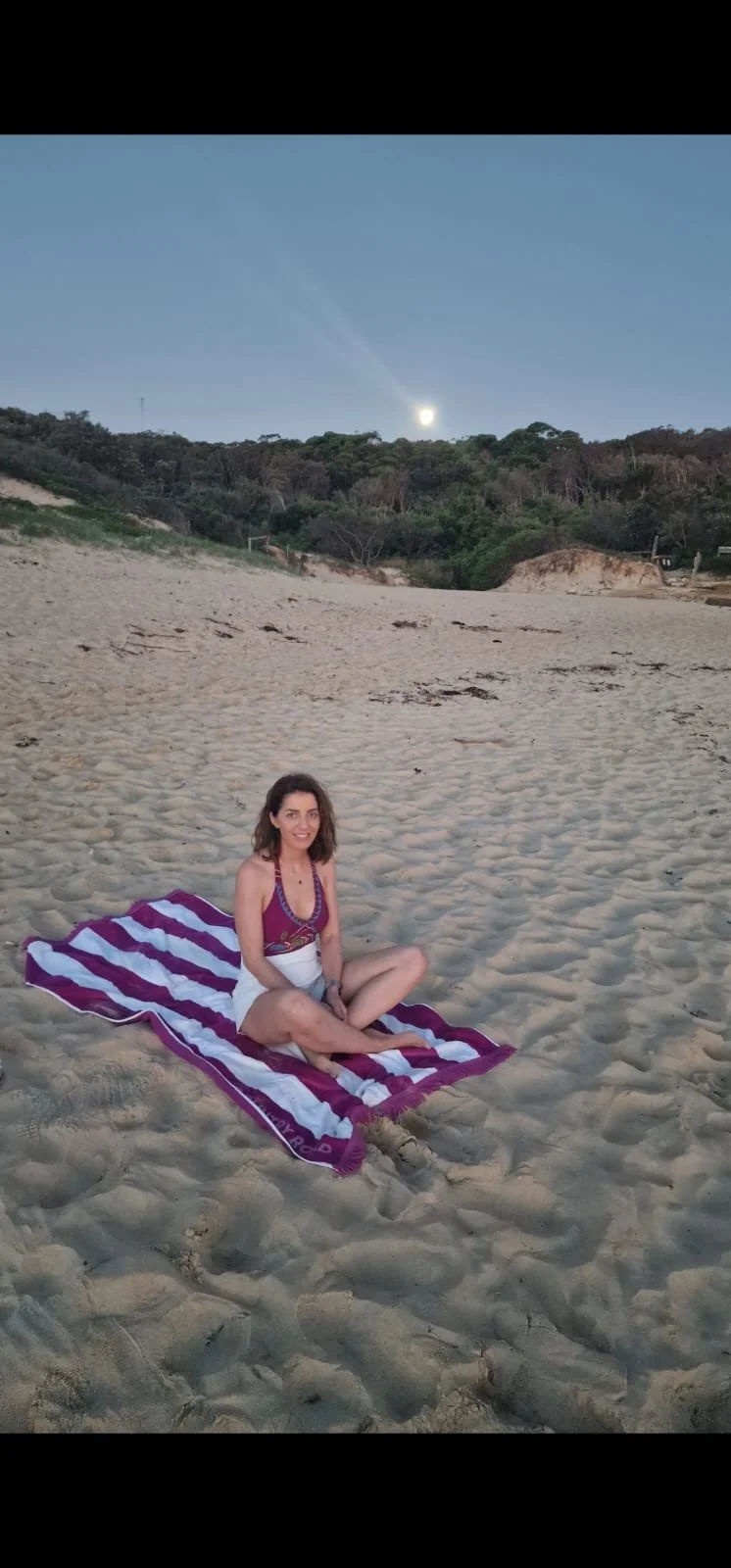 A woman sitting on a striped towel on a sandy beach at dusk, with a hill covered in trees and the moon visible in the sky.