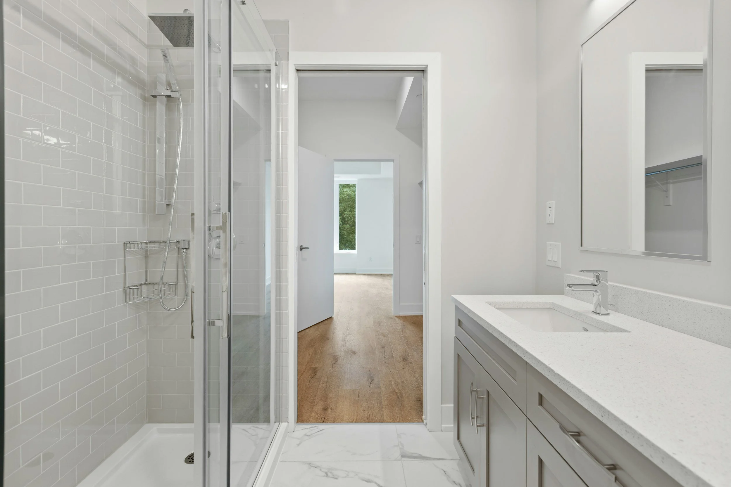 Baño moderno con ducha, lavabo y espejo, con vista hacia una habitación con suelo de madera y ventana
