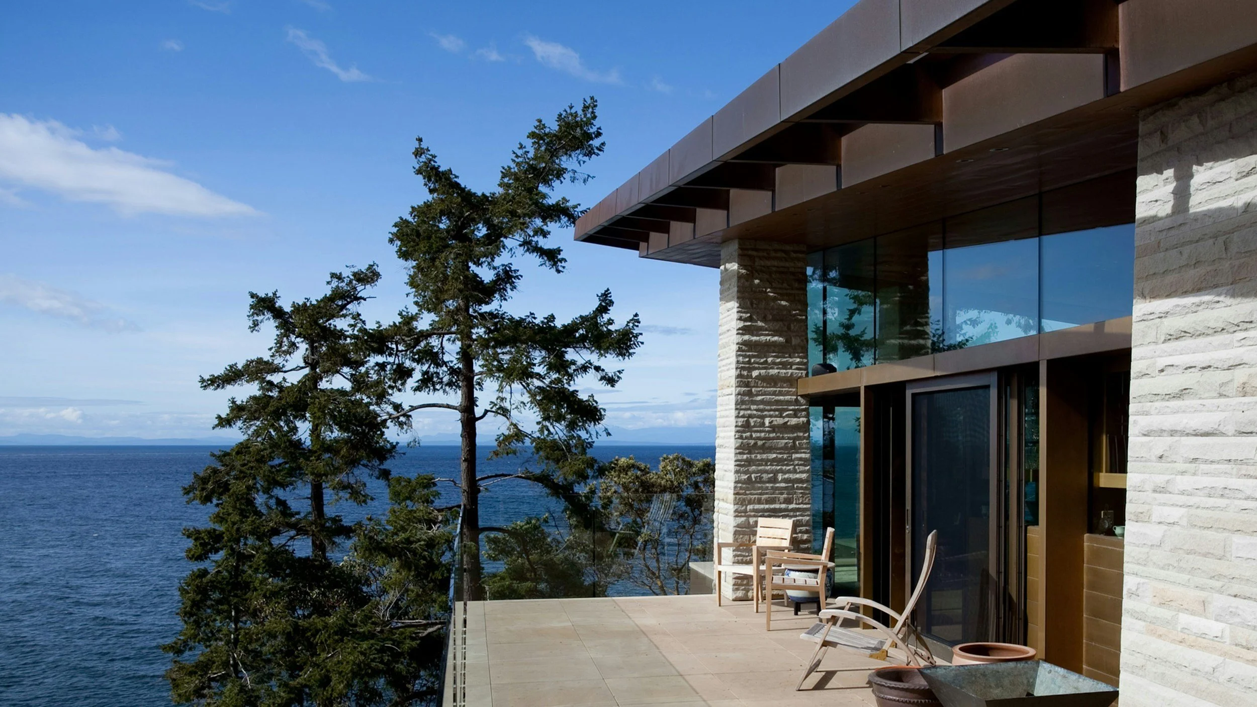 Terraza con sillones de madera, vista al océano y árboles cercanos, parte de una casa moderna con paredes de piedra y ventanas grandes.