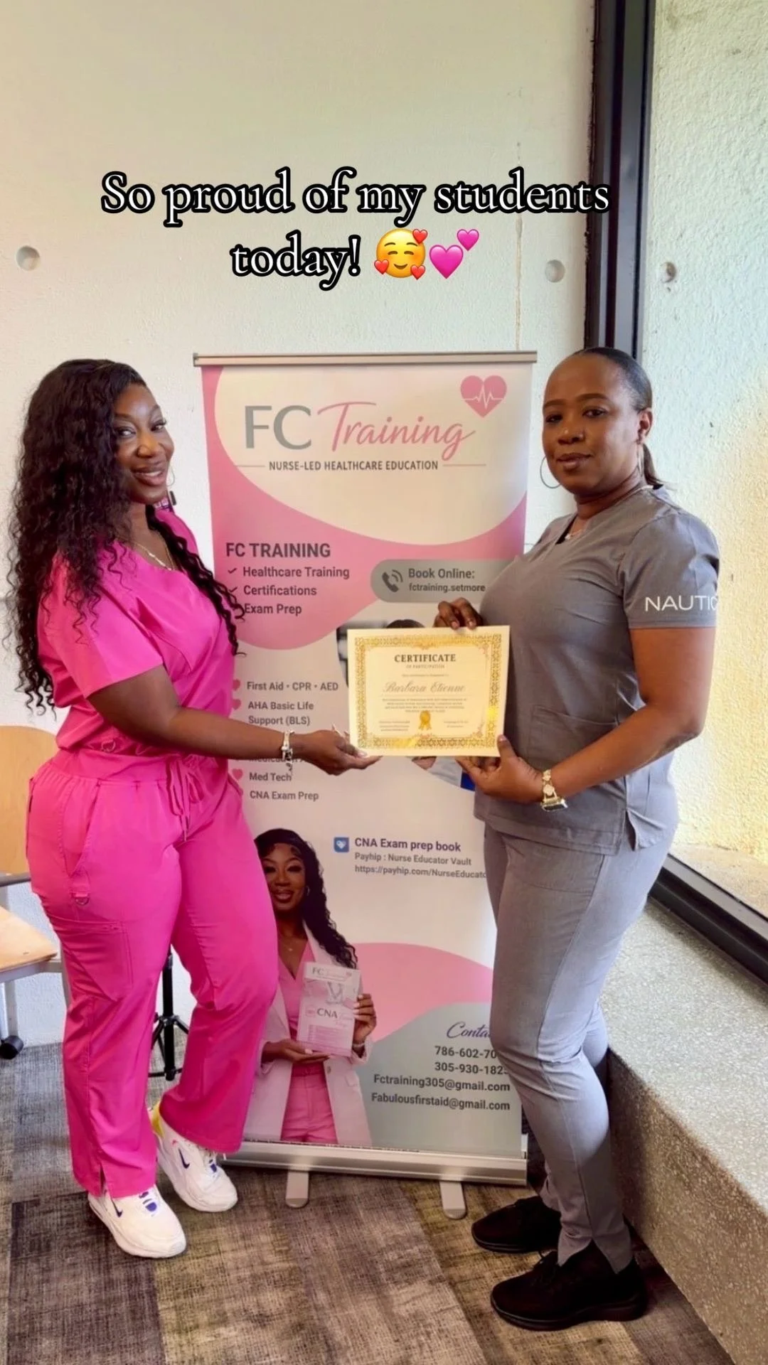 Two women are standing indoors, with one woman wearing pink scrubs and holding a certificate, and the other woman in gray holding the same certificate. They are standing in front of a pink and white banner that reads 'FC Training' and lists healthcare training programs. The woman in pink is smiling, and the woman in gray has a neutral expression.