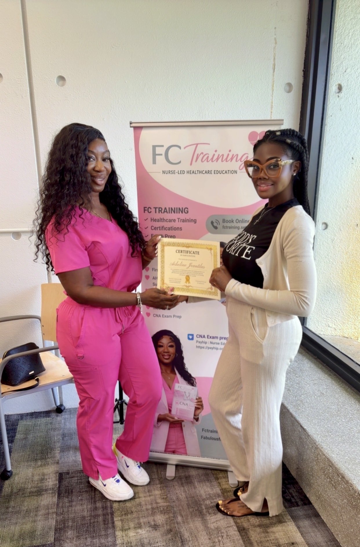 Two women standing indoors, one in pink medical scrubs and the other in beige pants and a black T-shirt, exchanging a certificate in front of a FC Training banner. The woman in pink is smiling at the camera, and the woman in black is wearing glasses and also smiling.