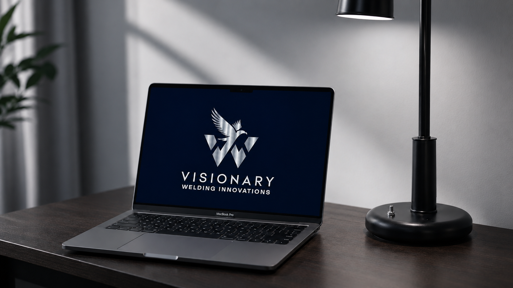 A laptop on a wooden desk displaying the Visionary Welding Innovations logo, with a black desk lamp nearby.