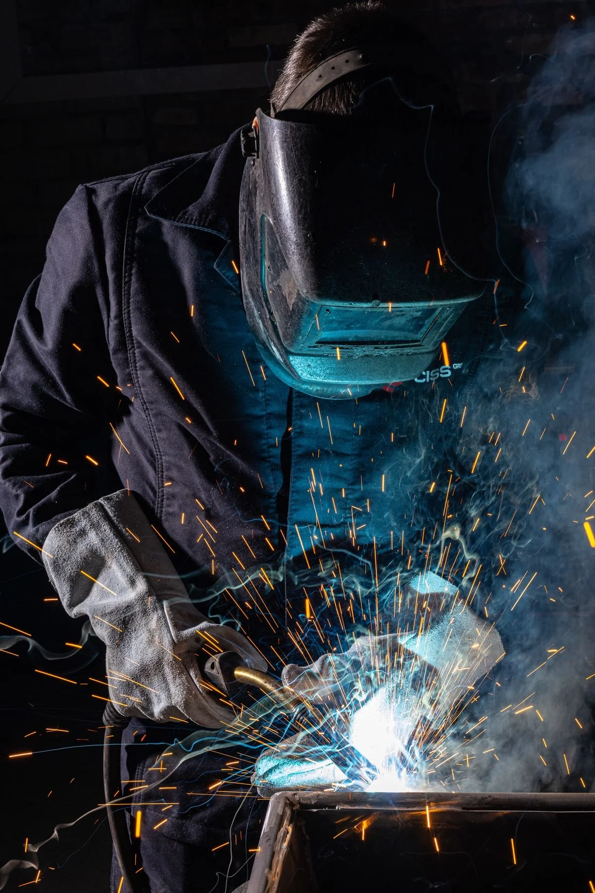 A welder in protective gear welding metal, sparks and smoke flying in the workshop.