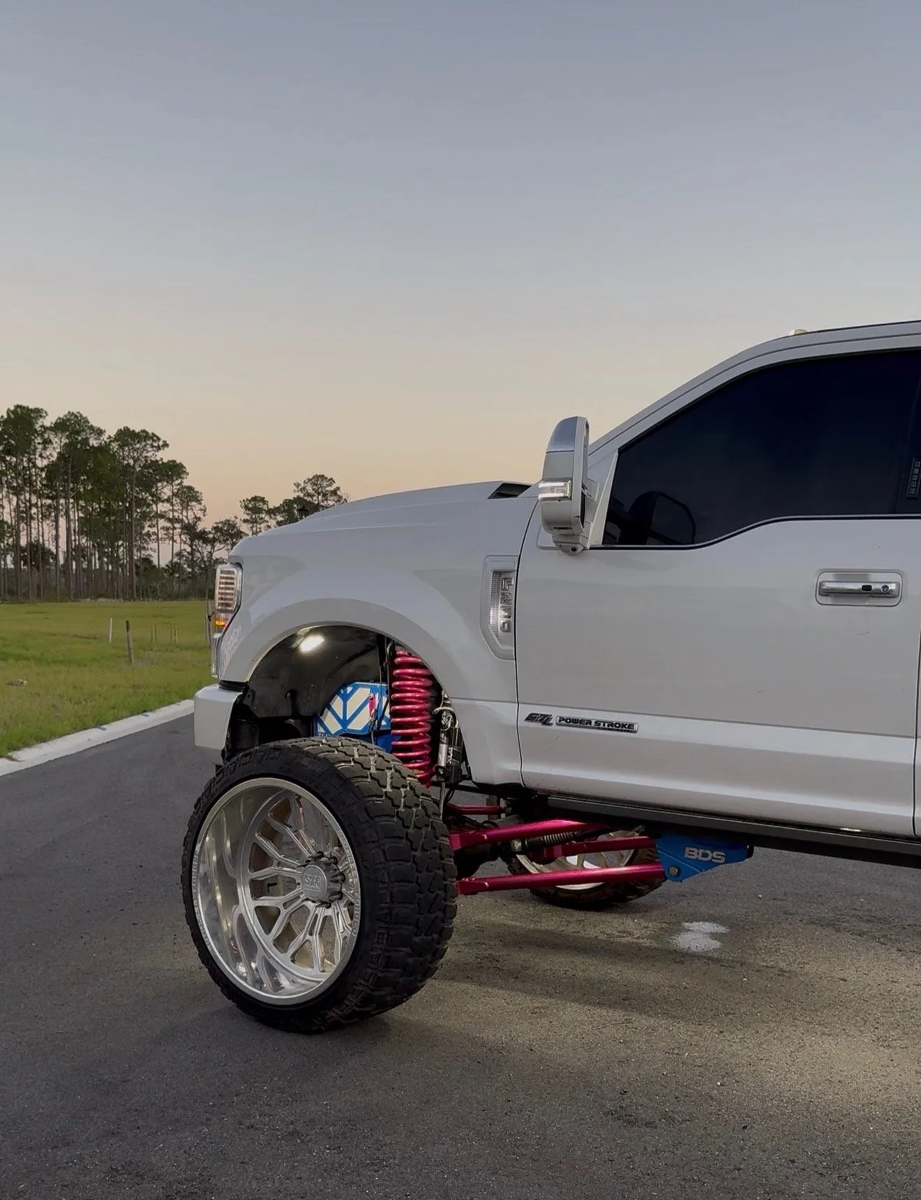 Partially assembled white off-road truck with large tires and red suspension components, parked on a paved surface at dusk with a background of trees and a clear sky.