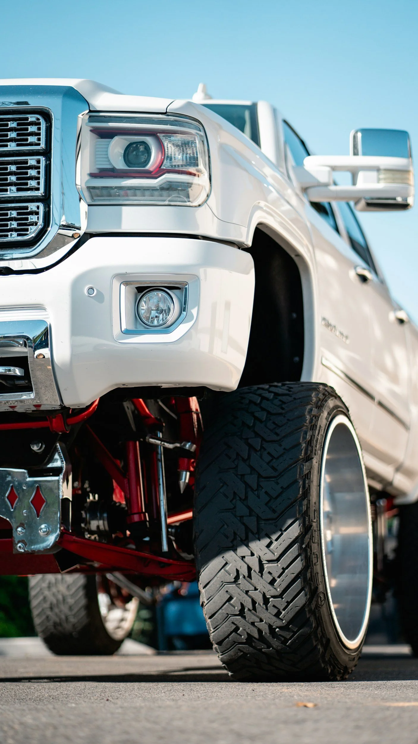 Close-up of a white lifted truck with large tires, showing the front driver side, with clear blue sky in the background.