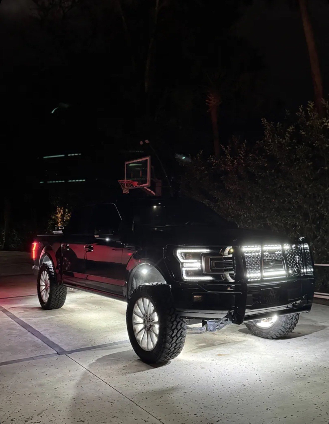 A black pickup truck with large tires parked on a well-lit parking lot at night, with a basketball hoop and trees in the background.