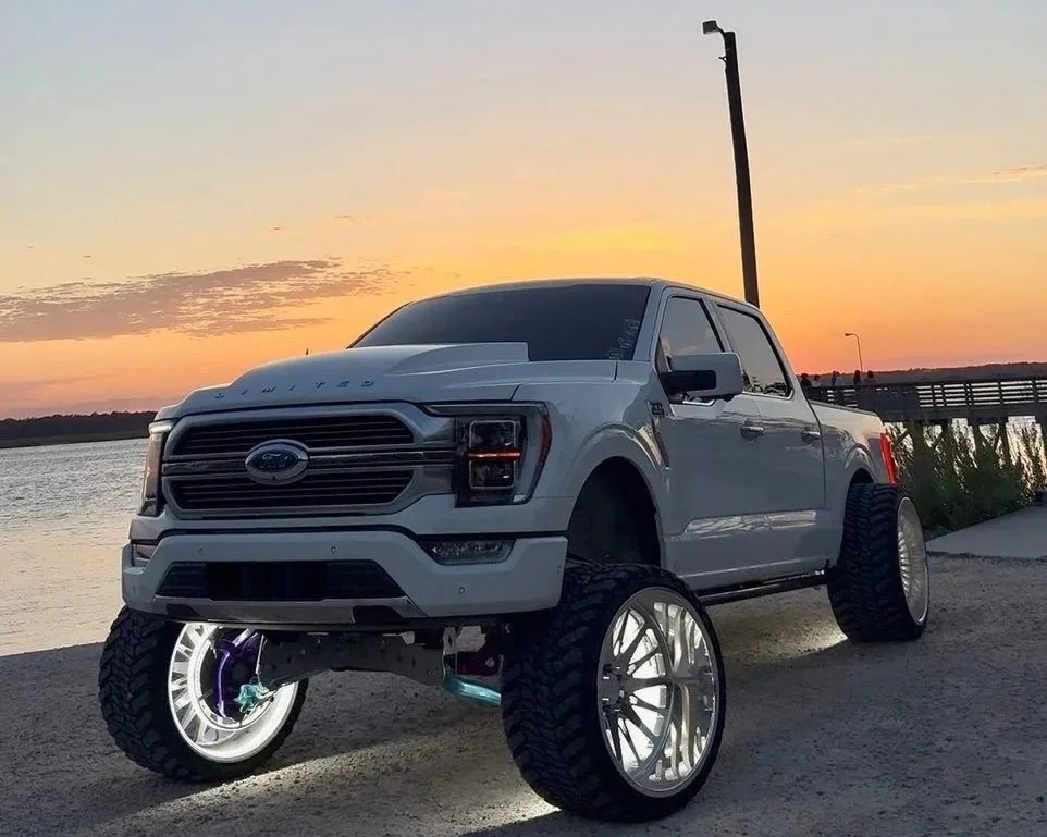 A white pickup truck with oversized, shiny silver wheels parked near a body of water during sunset.