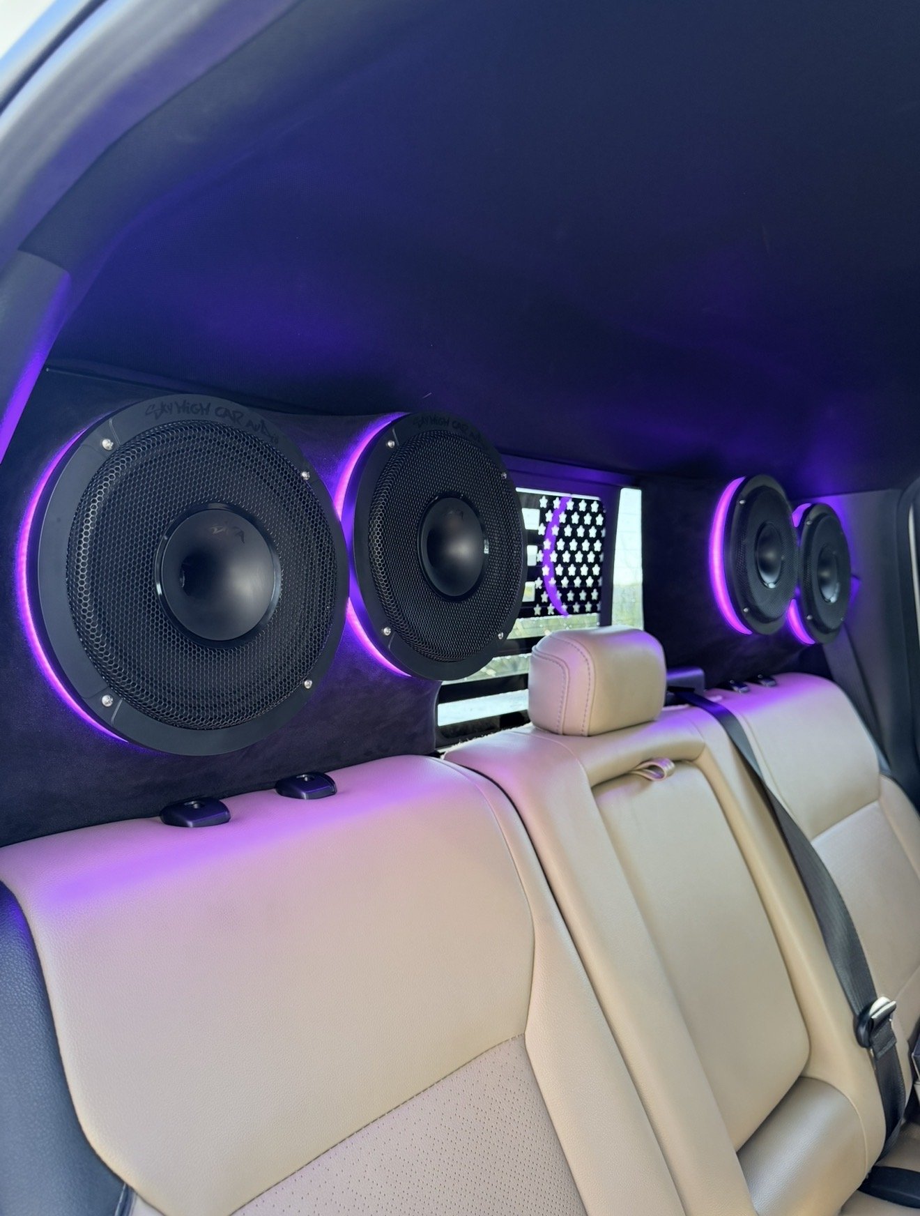 Inside the back of a vehicle with beige leather seats and a black ceiling, featuring four large black speakers with purple LED lighting and a small illuminated American flag.