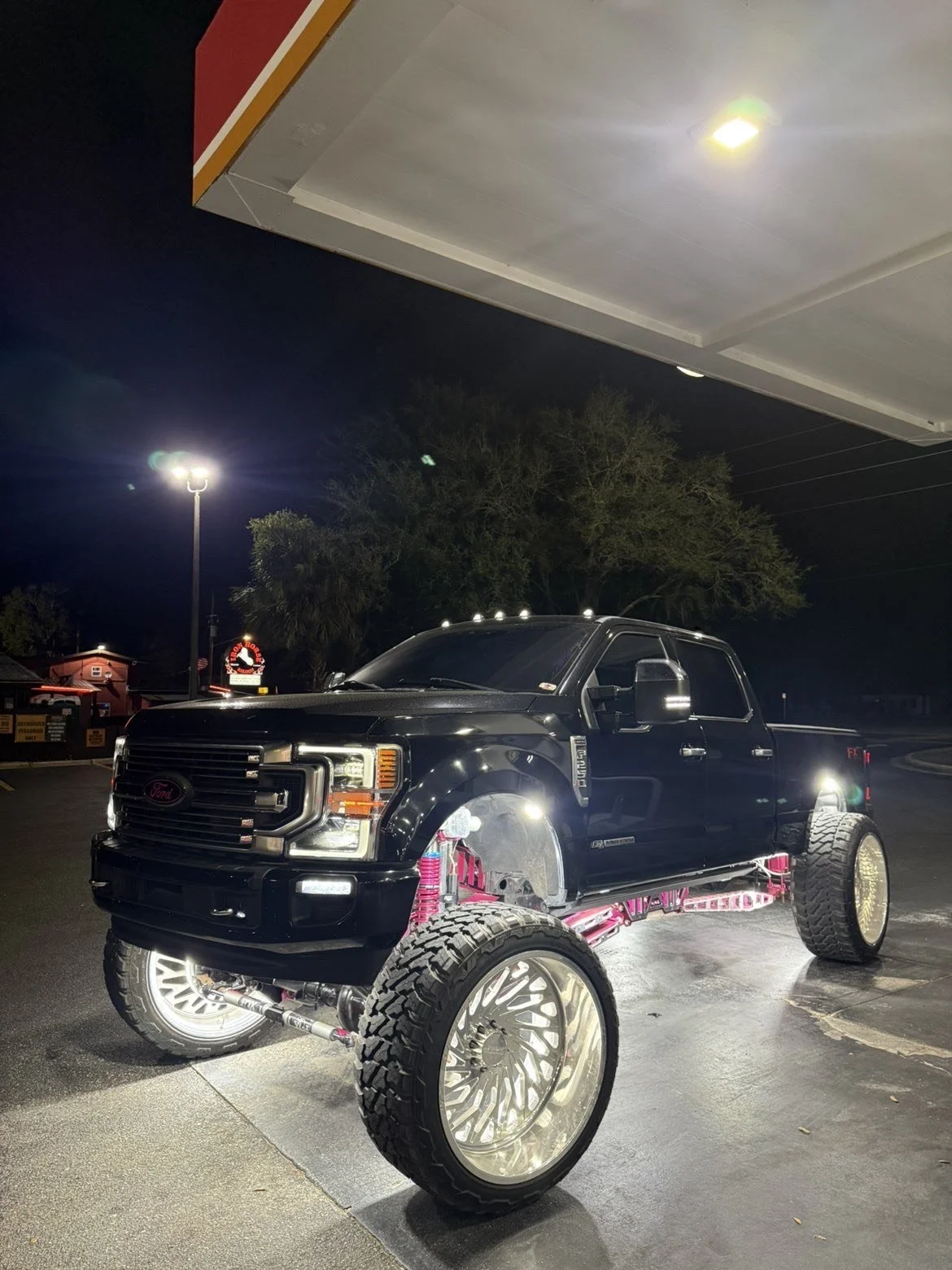 A black Ford pickup truck with custom wheels and pink suspension parts, parked in a lot at night under a canopy, with trees and a building in the background.