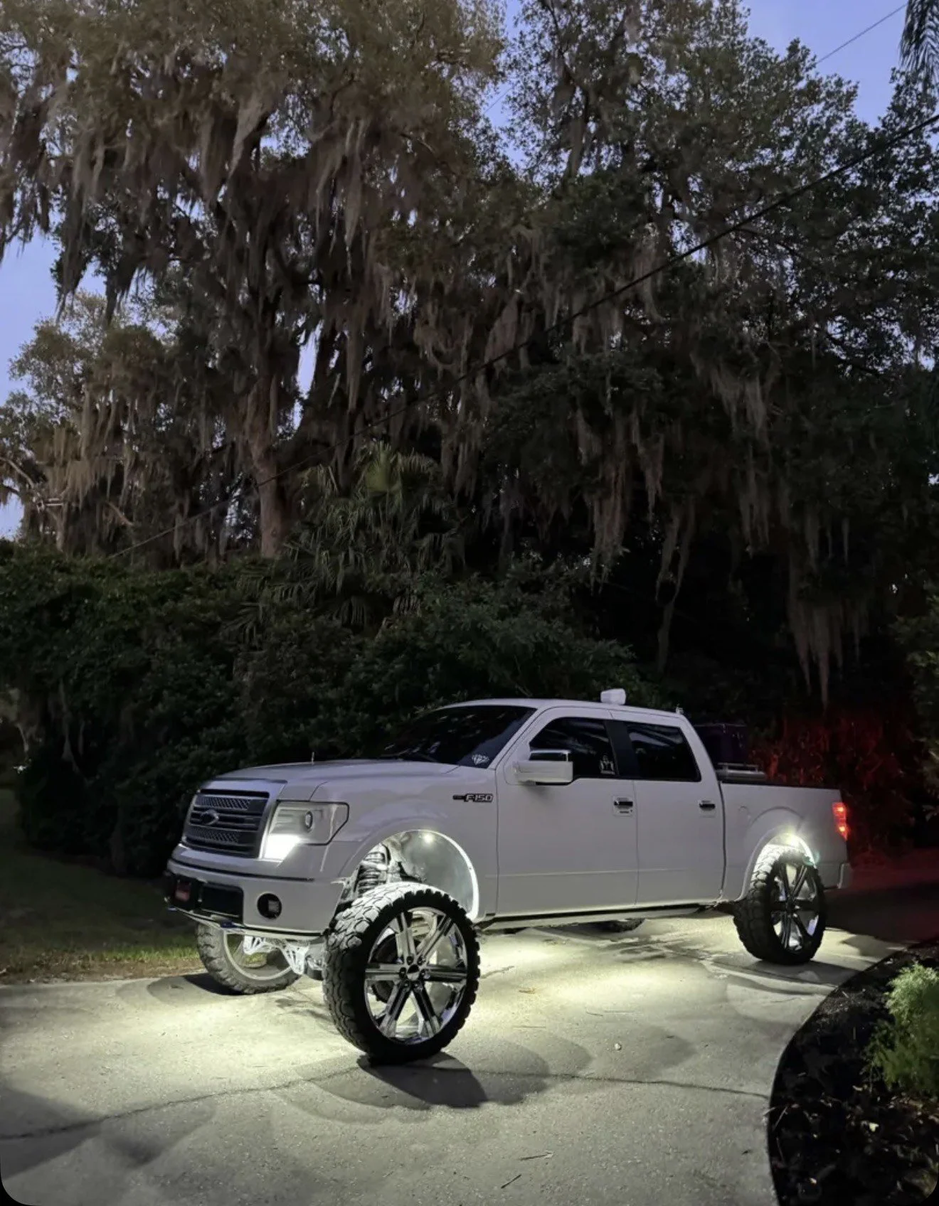 A white pickup truck with large, custom wheels is parked outdoors at night, under trees with Spanish moss. The truck's front and rear wheels are missing, revealing the suspension and axle.