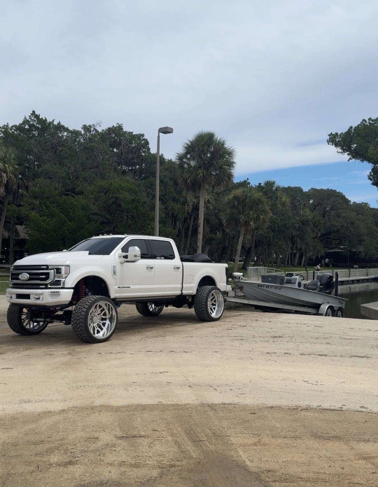 A white pickup truck with large custom wheels and lifted suspension parked on a sandy area near a small boat on a trailer, with trees, a lamppost, and a blue sky in the background.