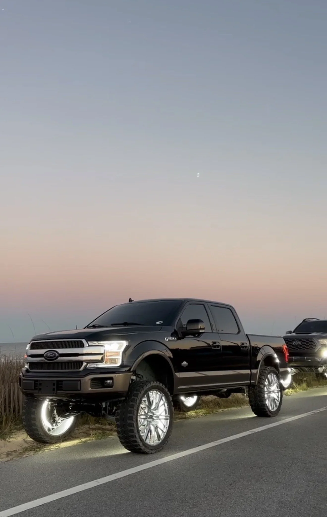 A black Ford pickup truck with large, shiny wheels and off-road tires parked by the side of the road during sunset, with the sky transitioning from pink to blue.