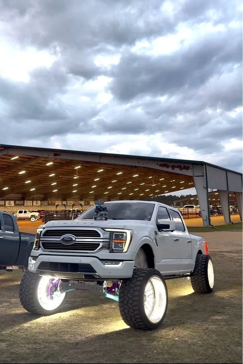 A silver Ford pickup truck with oversized wheels and illuminated underglow lights parked outdoors near a covered area, with cloudy skies above.