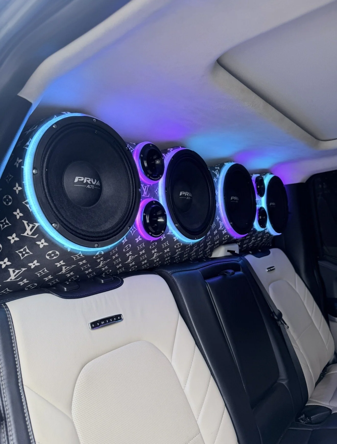 Car interior with a sound system featuring four large speakers with colorful LED lights and a Louis Vuitton patterned cover, installed along the back of the front seats.