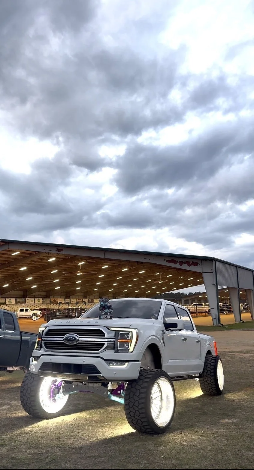 A modified white Ford pickup truck with large off-road tires and illuminated white rims, parked outdoors near a covered area with other vehicles in the background under a cloudy sky.