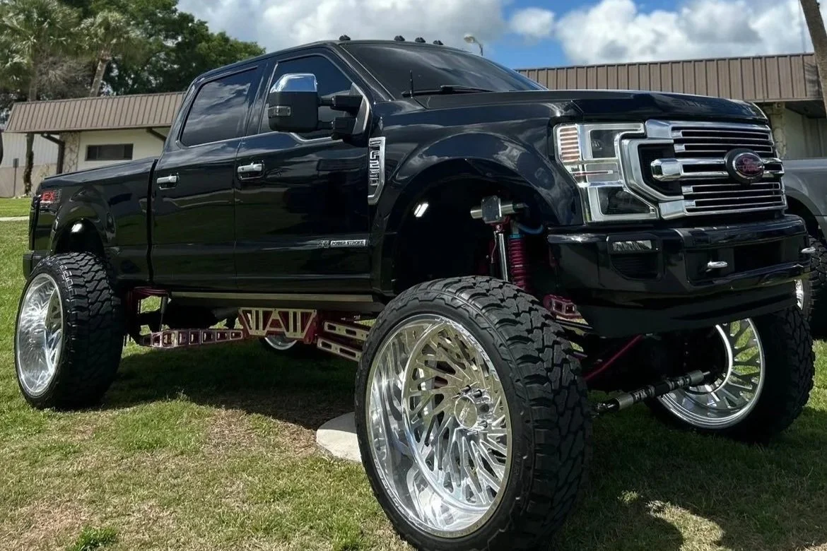 Black lifted Ford truck with oversized chrome wheels and exposed suspension components parked on grass.