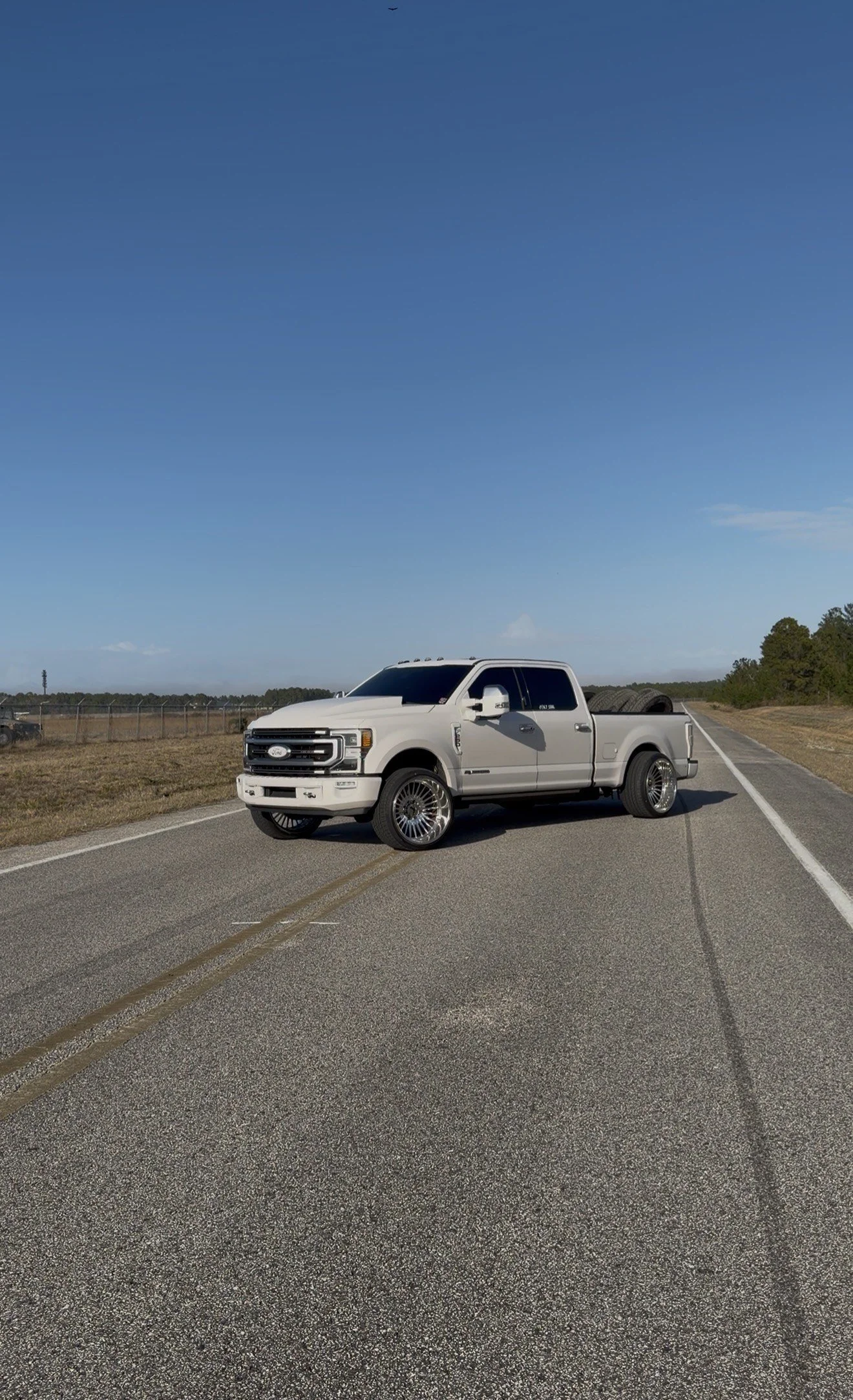 White pickup truck parked on an empty road with a clear blue sky above and trees on the horizon.