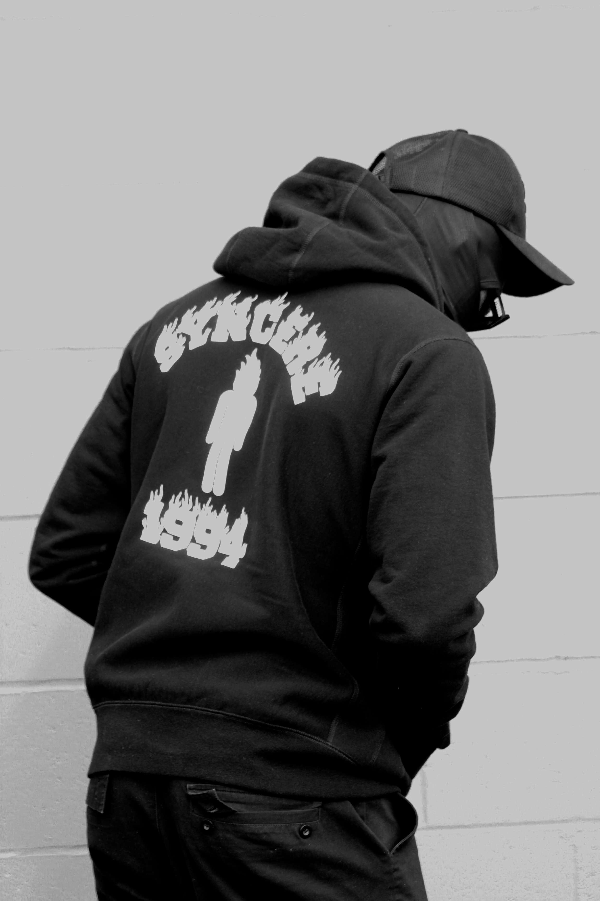 Person wearing a cap and a hoodie with graphic text on the back, standing against a plain wall.